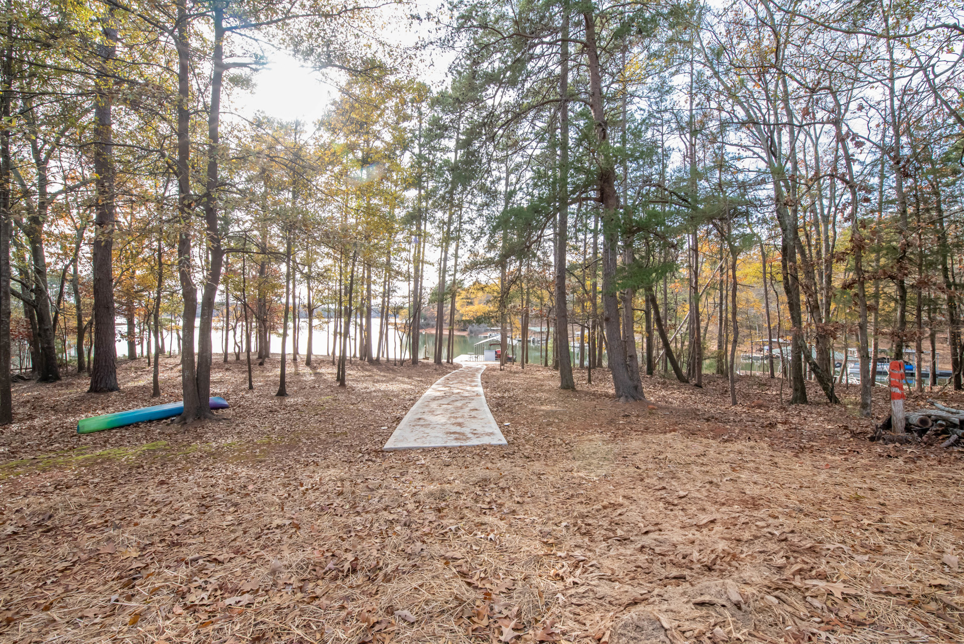 Winding dirt path through dense autumn forest with dry leaves covering the ground, tall deciduous trees, and a blue canoe resting near a lake