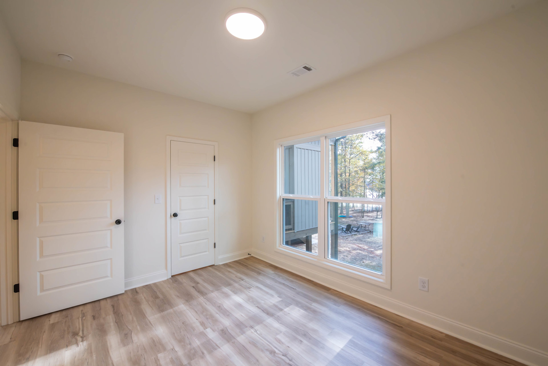 White walls, wood laminate flooring, ceiling light fixture, white door with black knob, window overlooking backyard.