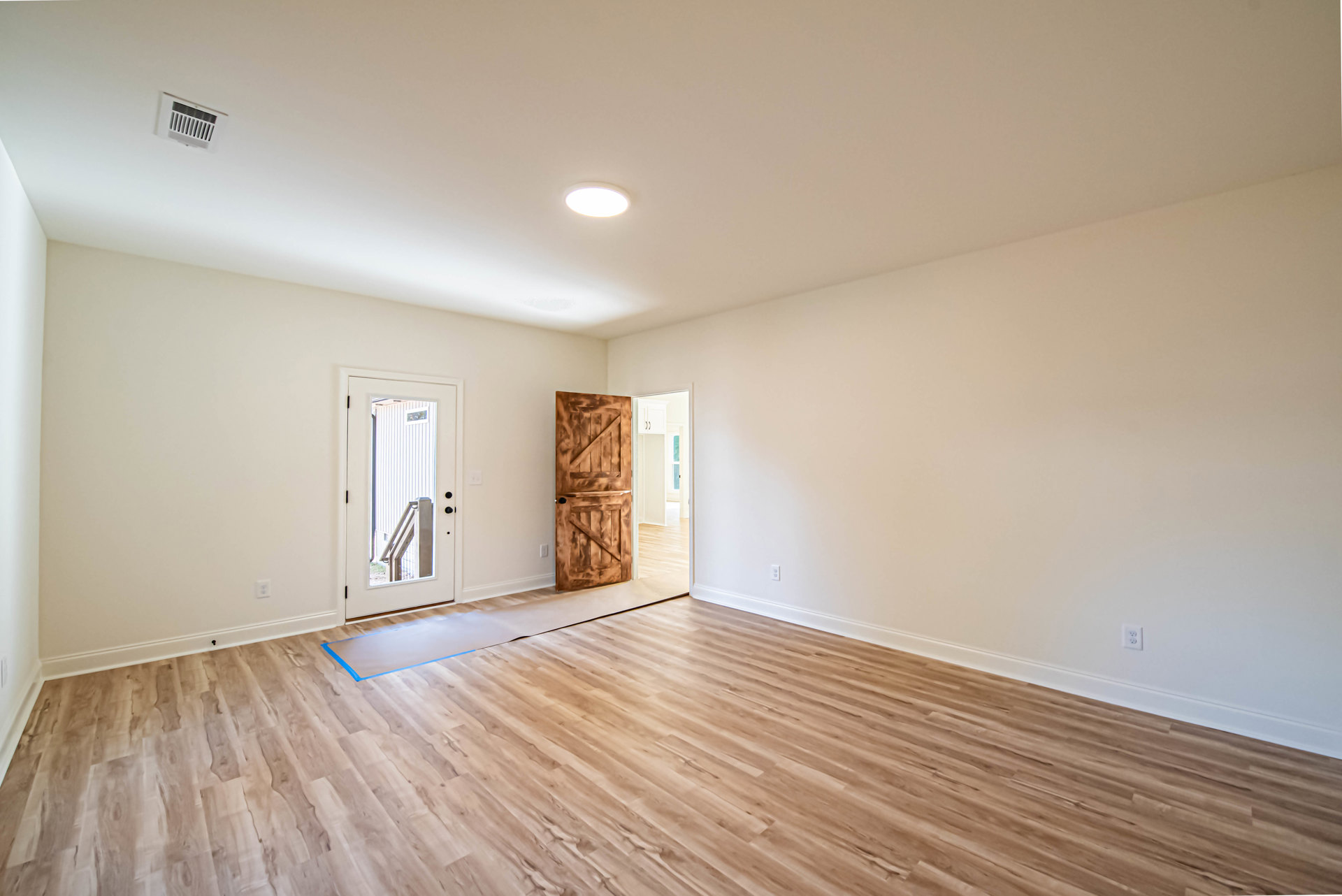 Wood flooring in a room with a wooden door featuring a black handle, metal railing, ceiling light, and ceiling vent.