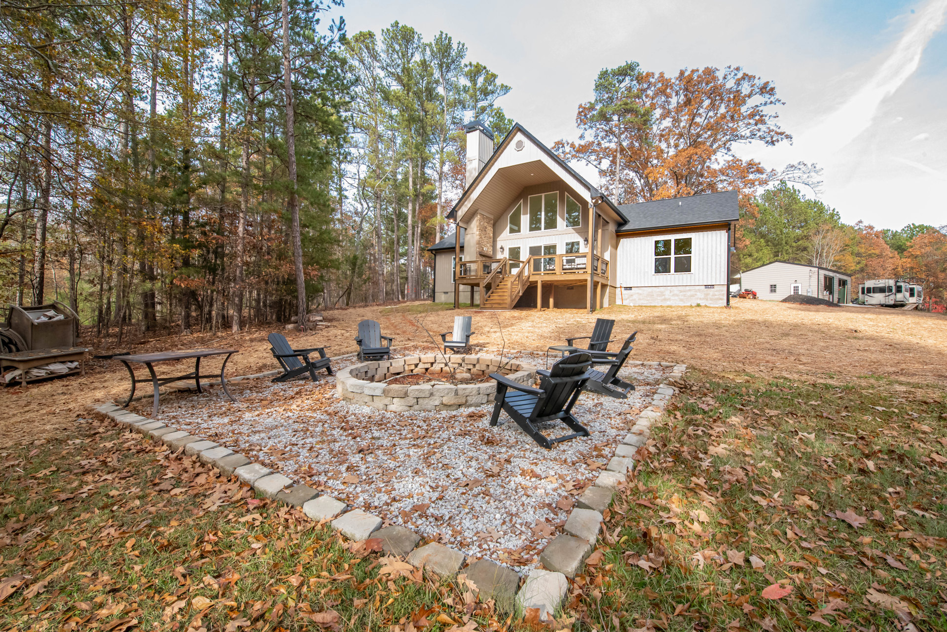 Modern house with wood siding, elevated deck, and stairs, surrounded by autumn leaves; gravel patio features a circular fire pit with black chairs arranged around it, trees and