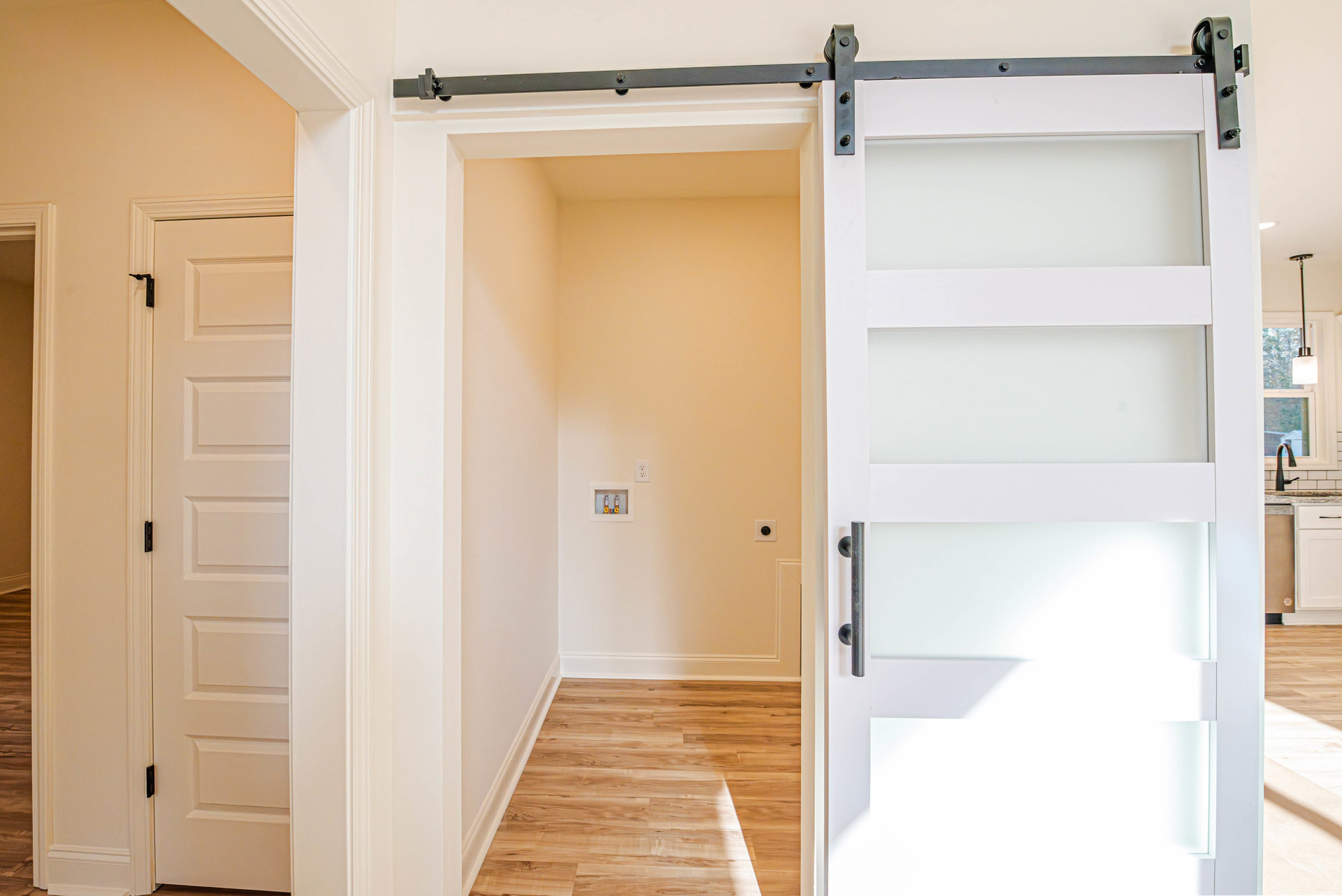 White hinged door with black handle beside sliding barn door, wood floor with white baseboard, black ceiling track, white walls