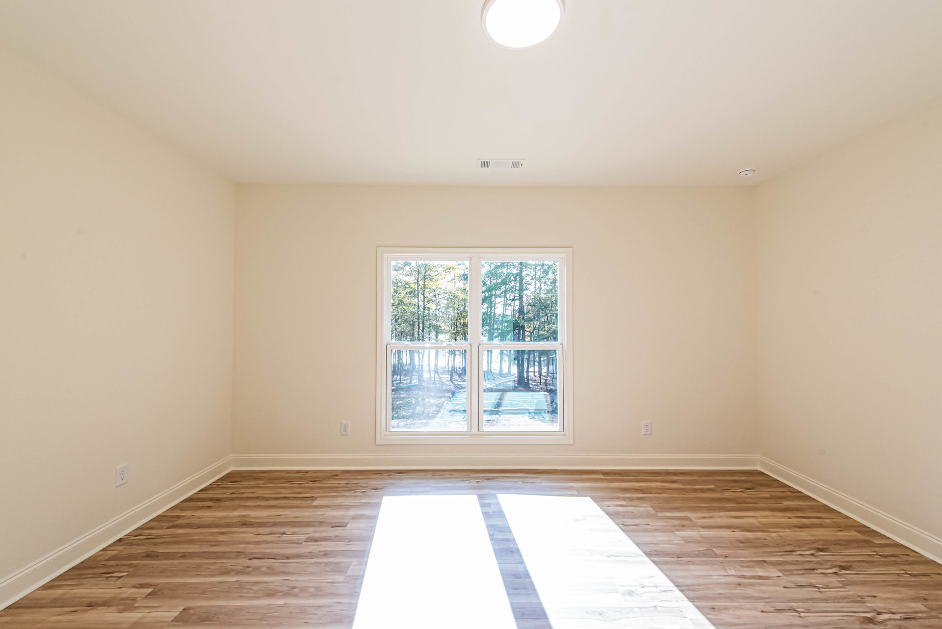 Sunlit room with wide window overlooking trees, wood plank flooring, white walls, ceiling light fixture, and minimalist decor.