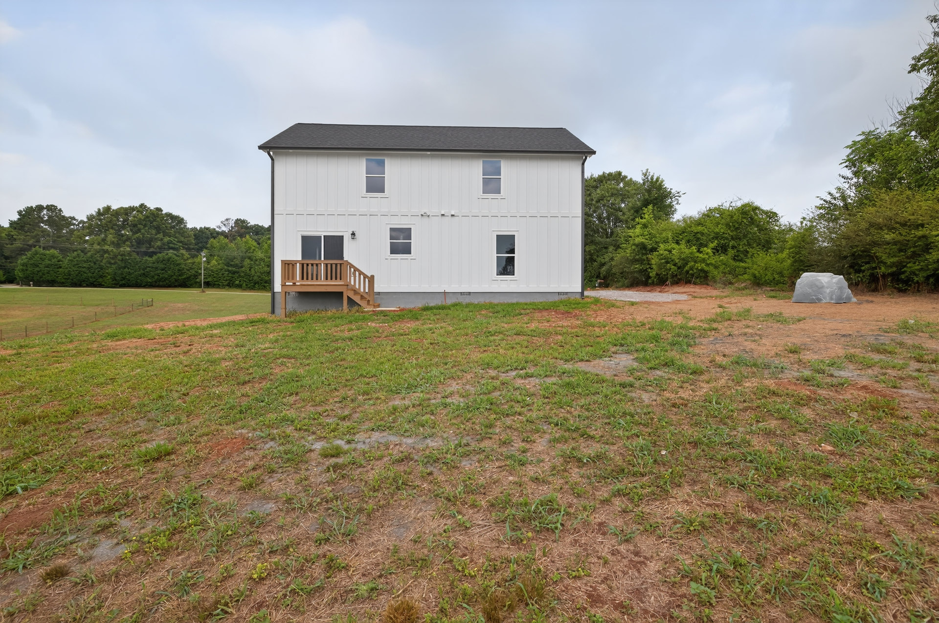 White house with large windows, wooden deck and staircase, wooden bench on grassy lawn, trees and cloudy sky in background