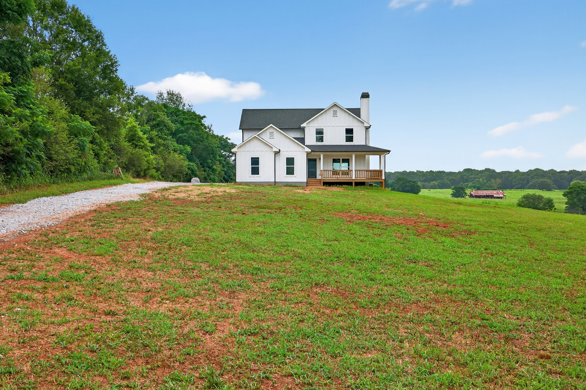 White farmhouse with chimney on grassy hill, wraparound porch with railing, wooden deck, gravel path bordered by trees and lawn under partly cloudy sky