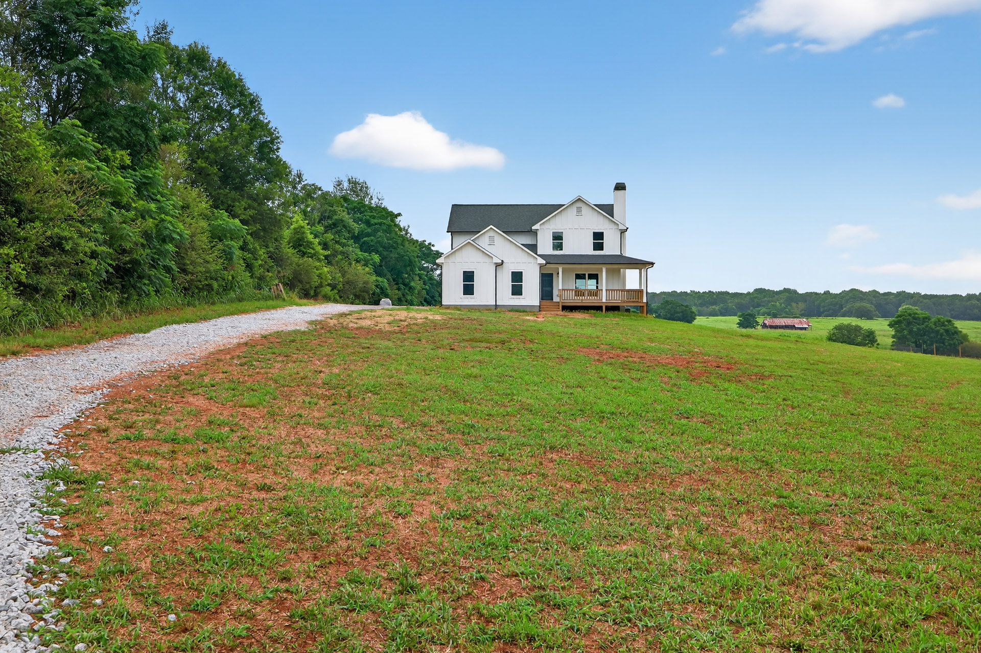 White house with black roof and large covered porch, situated on a grassy hill with trees and a wooden deck in the background, under a partly cloudy sky.