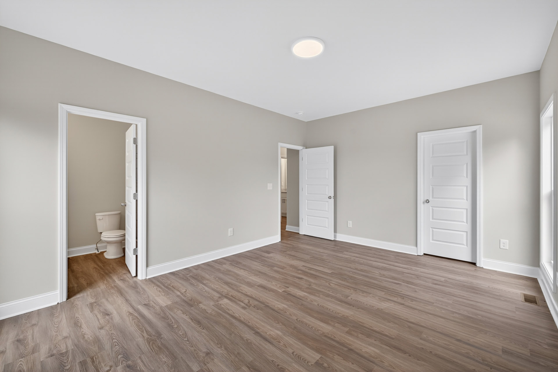 Room with light wood laminate flooring, white plaster walls, two white doors with silver hardware, and a toilet featuring a hose connected to the tank