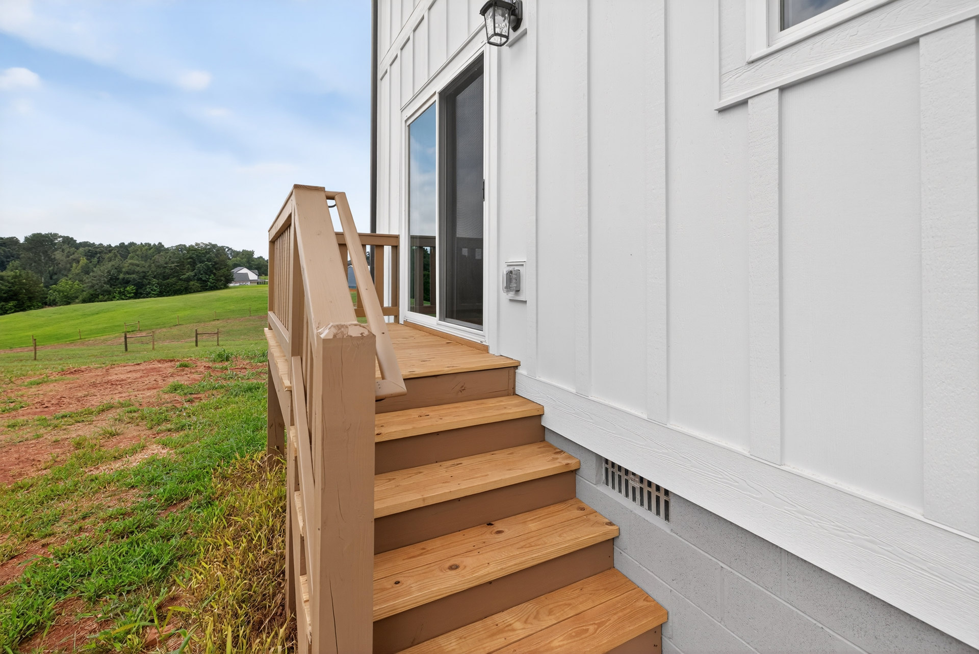 Wooden exterior staircase leading to porch, surrounded by grassy yard, trees, and wooden fence; large window visible on house facade.