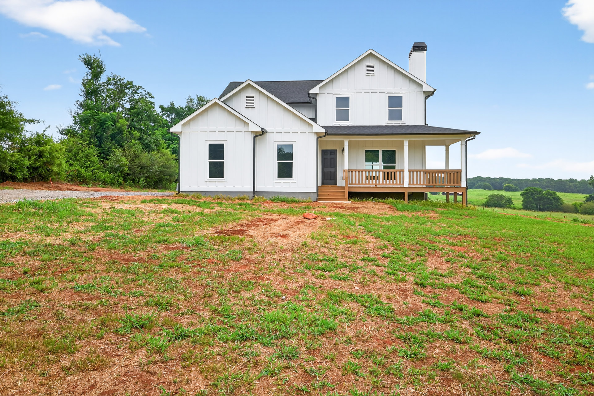 White farmhouse with covered porch, white framed windows, grassy lawn in foreground, trees and blue sky with clouds in background