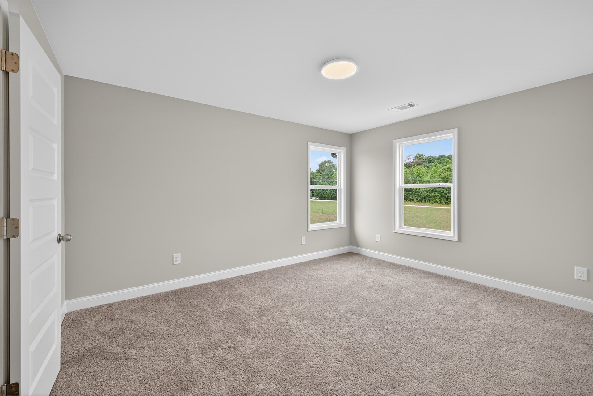 Bright carpeted room with large windows, white walls, ceiling light fixture, white paneled door, and view of green trees outside