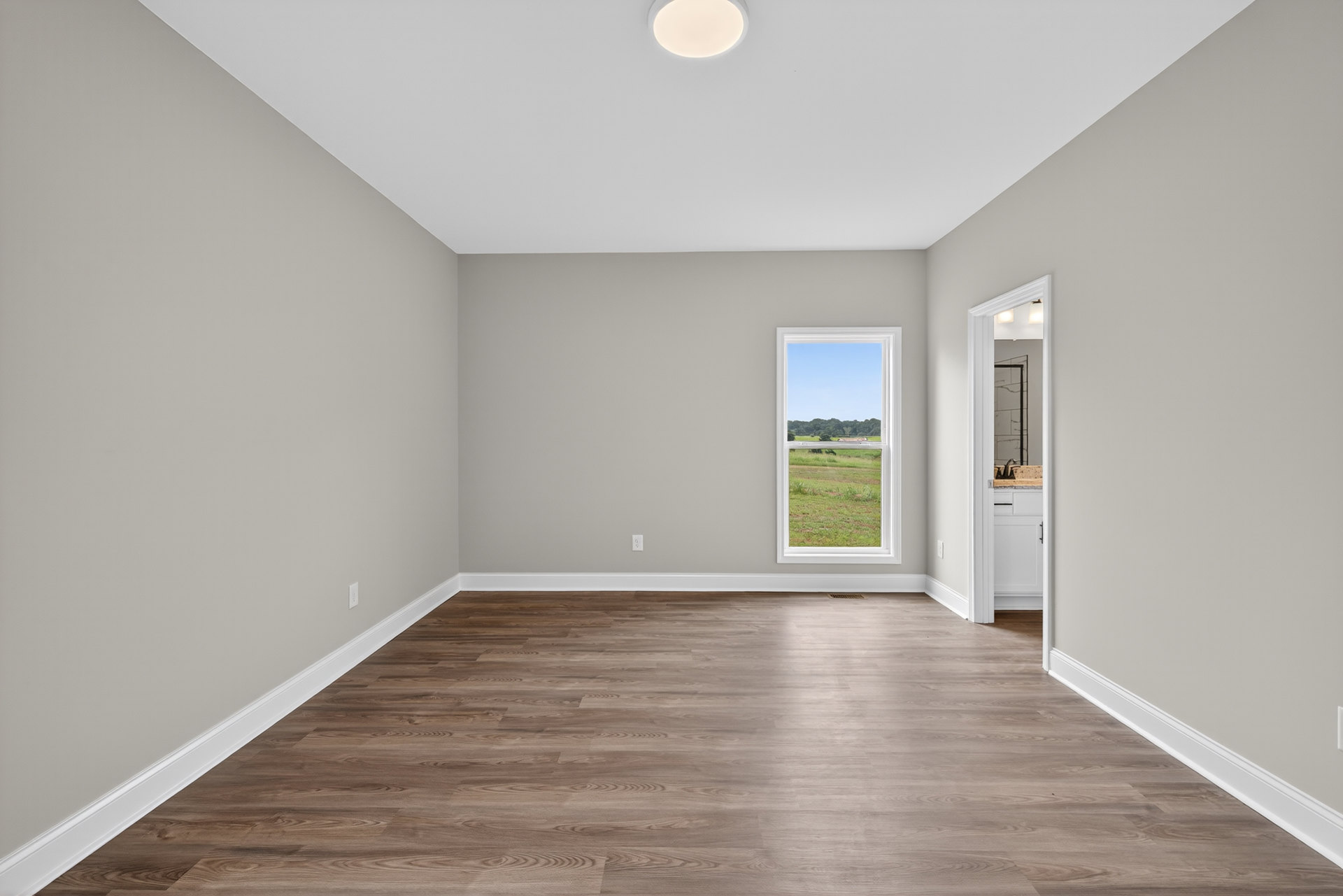 Sunlit room with wide wood plank flooring, white walls, large window overlooking grassy field, and white door with glass inset