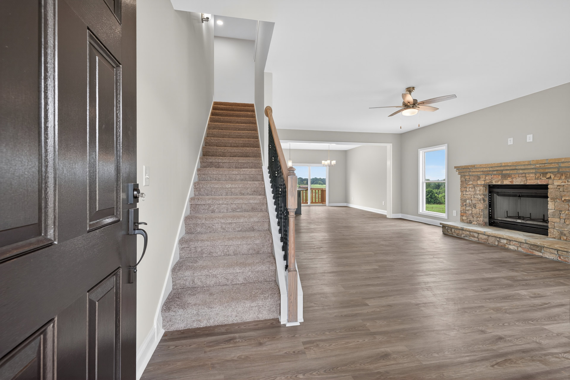 Living room featuring hardwood floors, white fireplace with glass door, wood staircase with decorative post, large window overlooking trees.