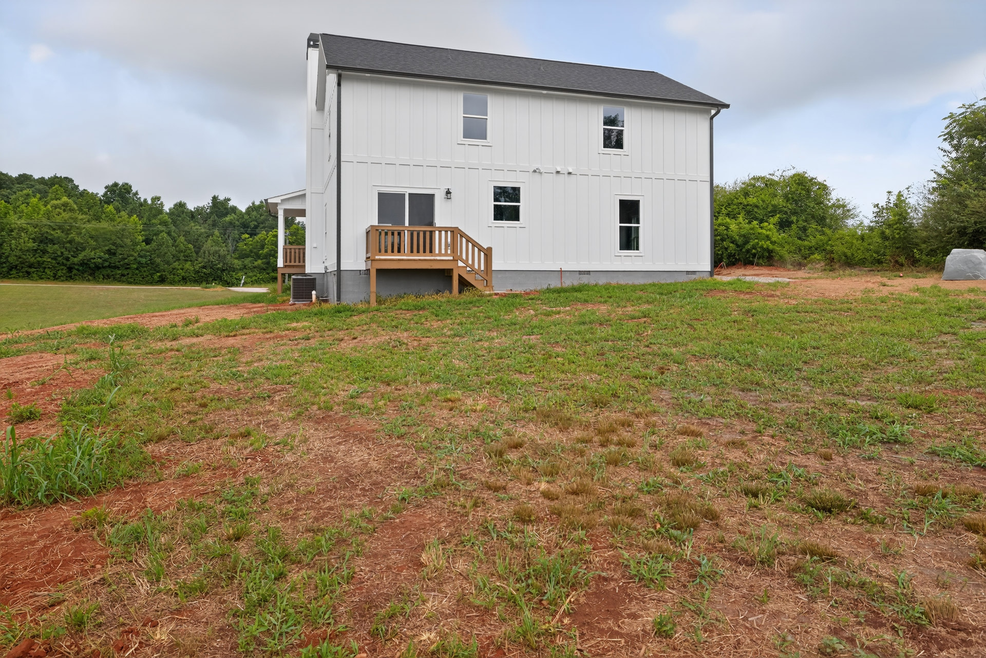 White farmhouse with black roof, wooden porch with railing, large windows, green lawn, and scattered trees under partly cloudy sky
