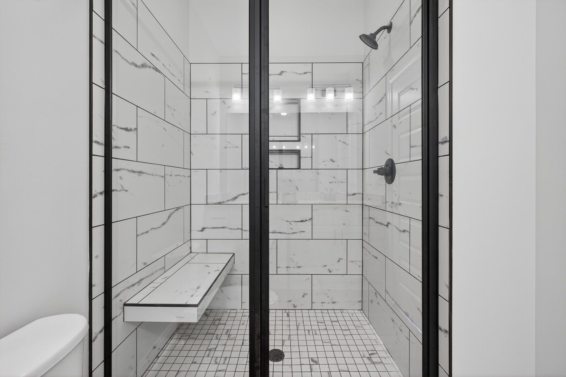 Walk-in shower featuring a white tile bench with black trim, matching tile floor with central drain, wall-mounted shower head, and partial view of a toilet and mirror.