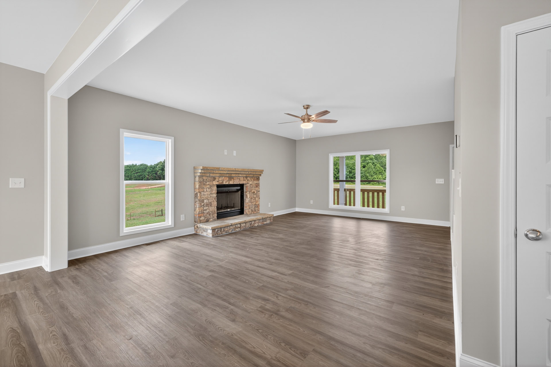 Spacious room featuring hardwood floors, white walls, stone fireplace, large window overlooking trees and field, ceiling fan with light fixture