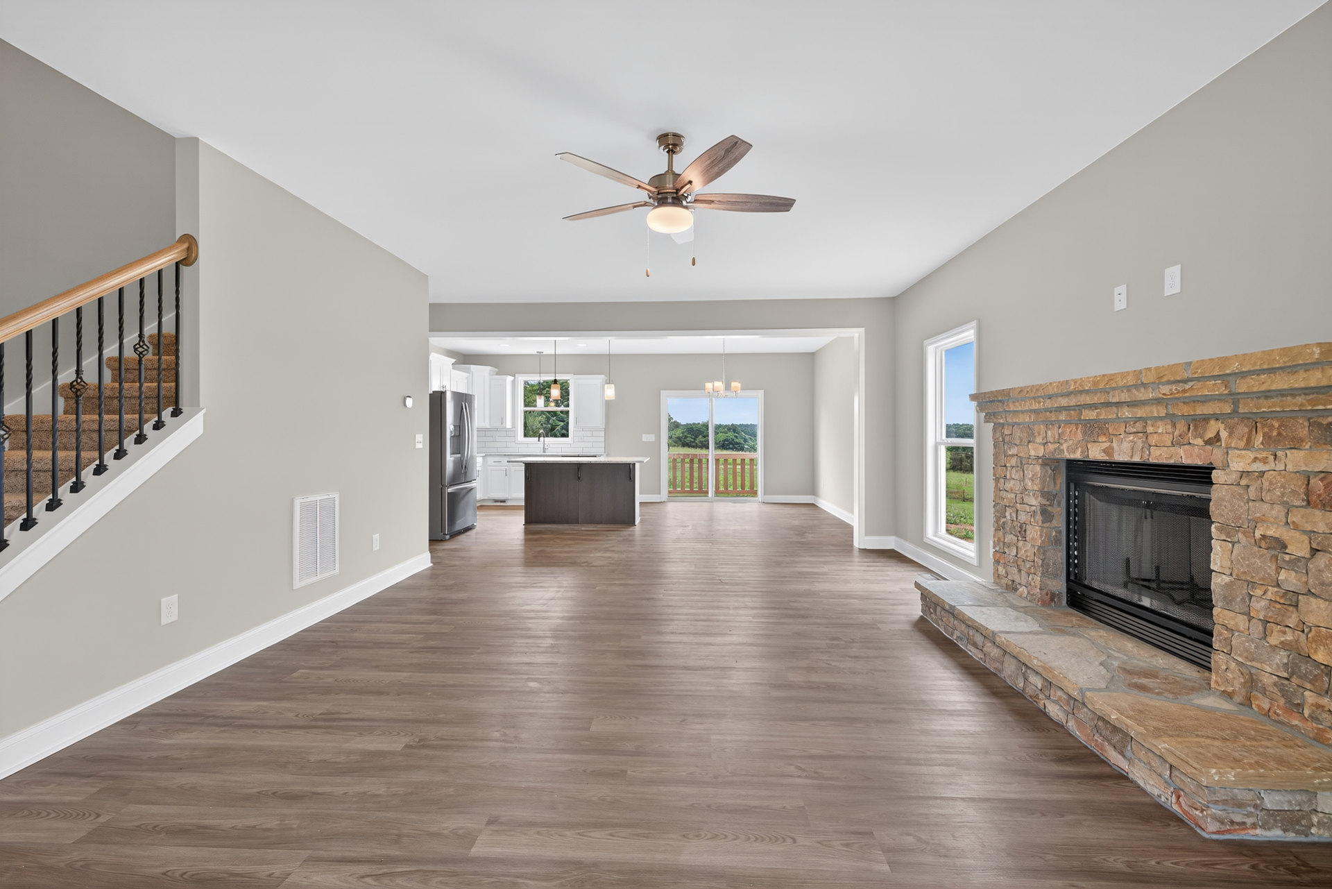 Living room with wood flooring, central fireplace with screen, ceiling fan with light, brown couch near staircase with black railing, large window overlooking field and trees
