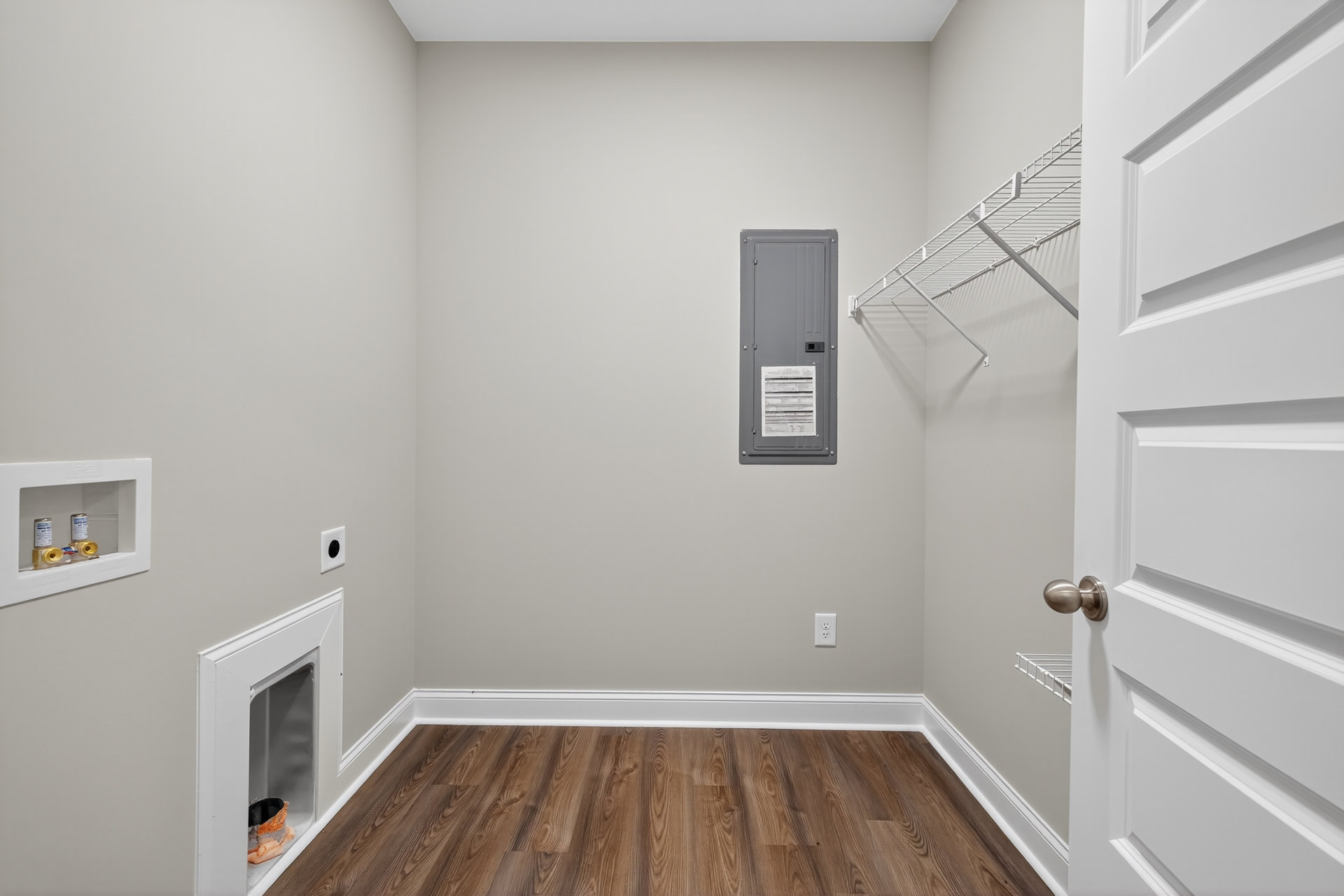 Wood floor with white trim, grey door featuring a vent, white rectangular object with a white frame, small white door containing a plastic bag of liquid, metal box against a