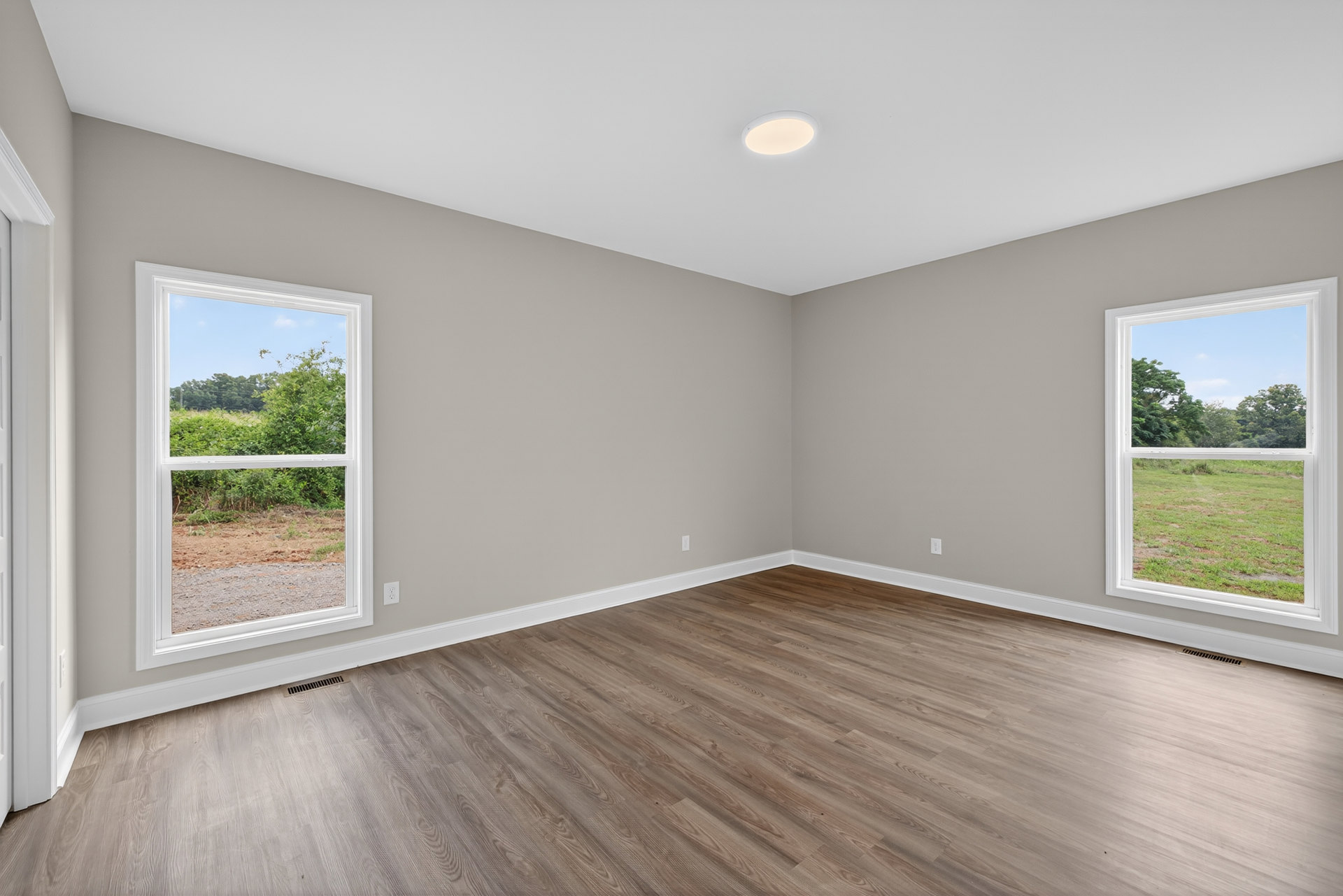 Sunlit room featuring wide-plank hardwood flooring, white walls, large window overlooking grassy field and trees, recessed ceiling light.