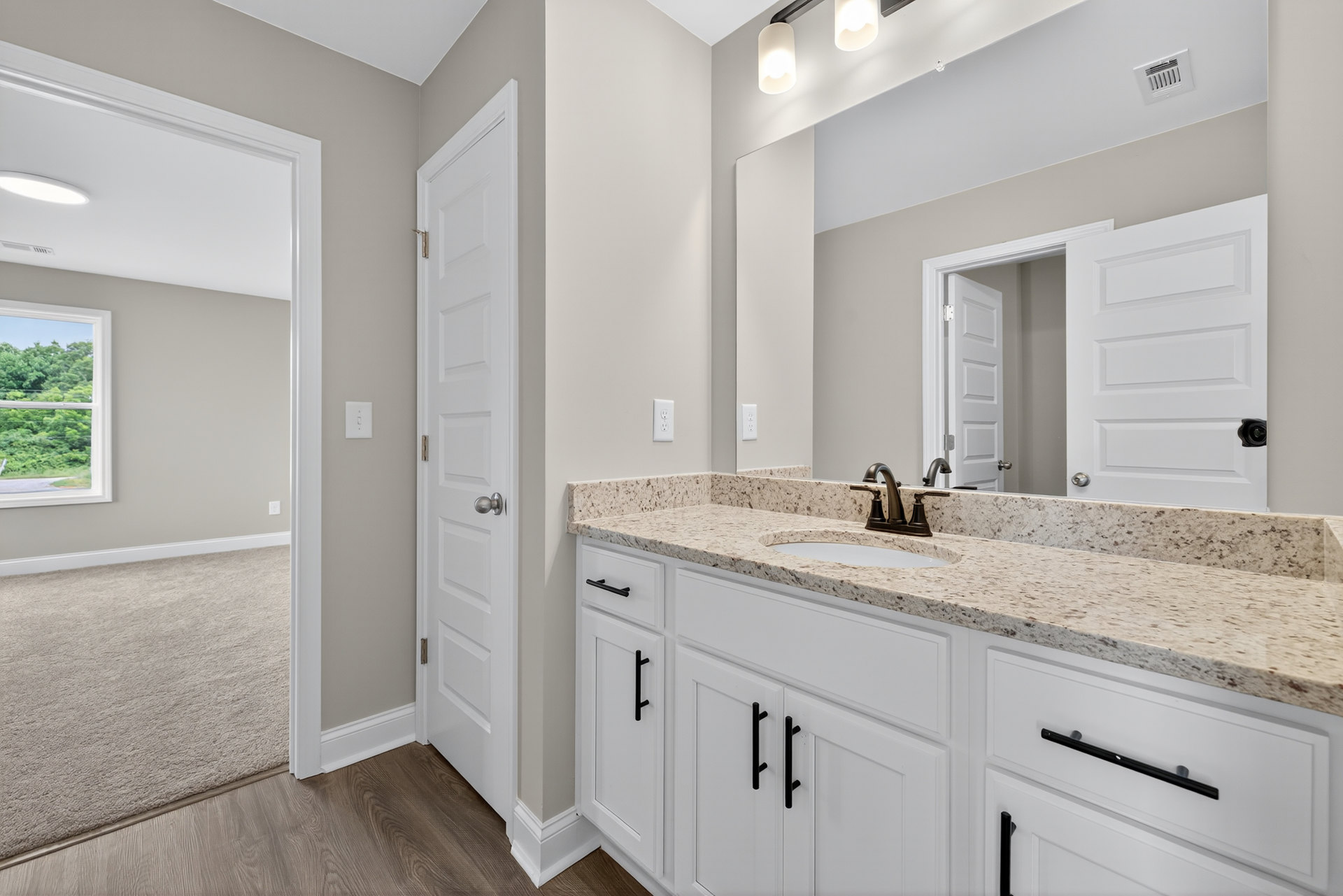 Bathroom with marble countertop, rectangular mirror above sink, white cabinetry, chrome faucet, window showing trees outside, wall vent, black pipe, and modern light fixture.