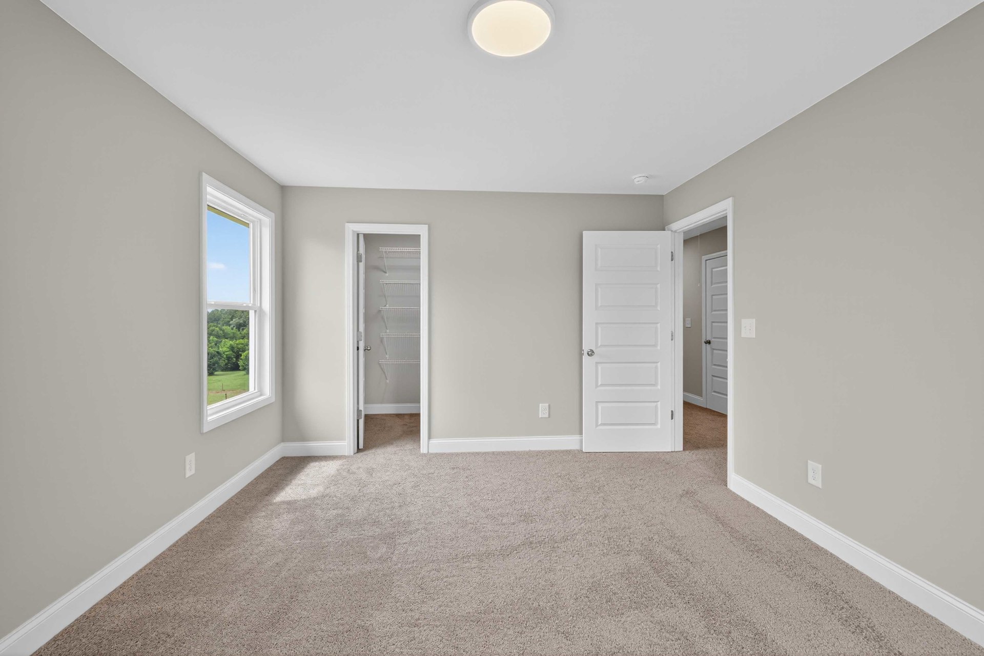 Beige carpeted floor, white paneled doors with silver handles, white walls, window overlooking green trees, simple baseboard molding