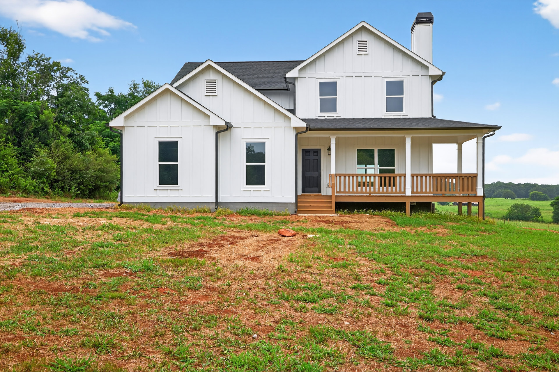 White siding house with black front door, covered porch supported by white columns, grassy lawn with patches of dirt, mature trees in background under blue sky with clouds