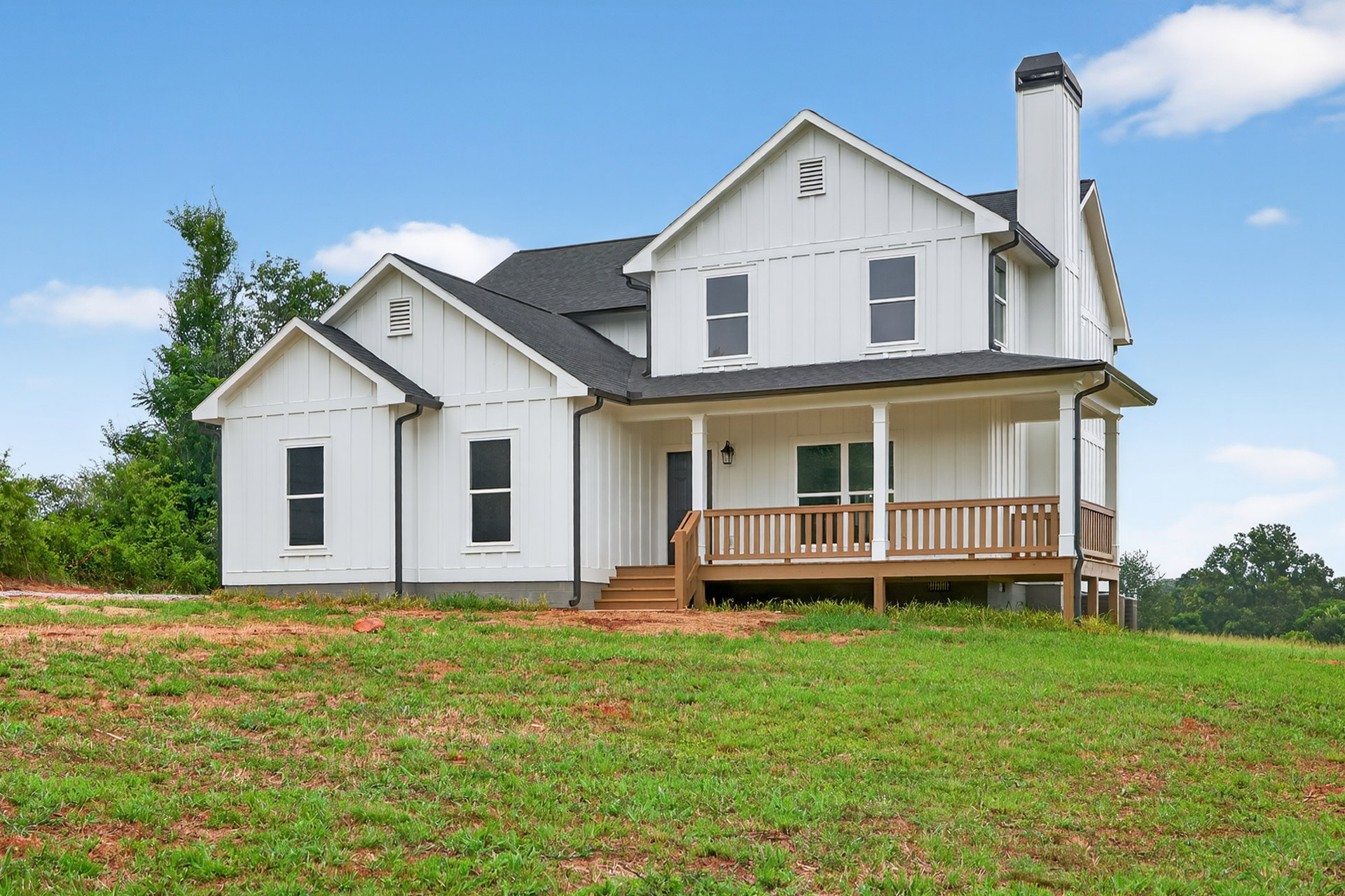White siding house with covered porch, wooden deck railing, grassy hill, and stairs leading up to entry; large windows and partly cloudy sky in background.