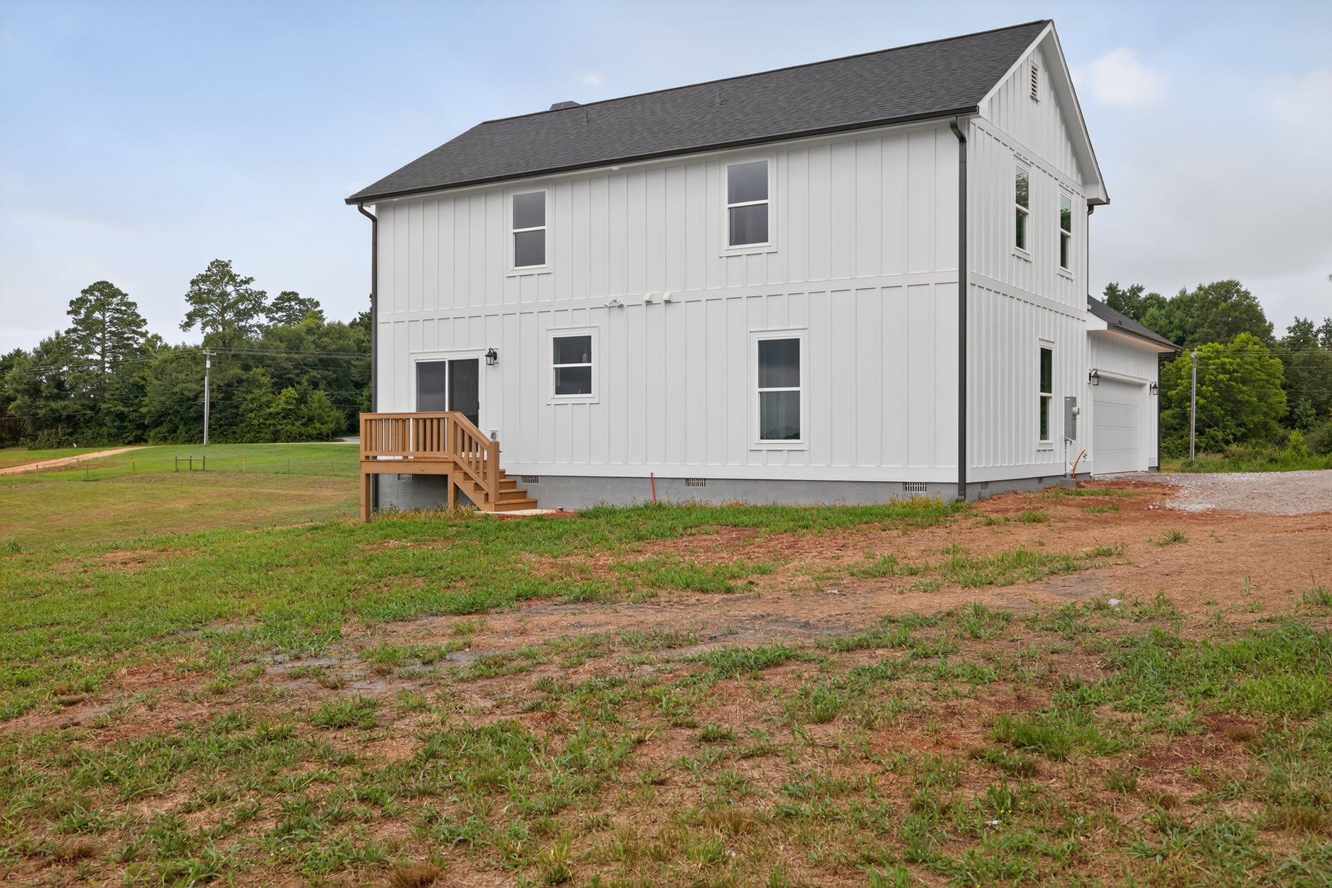 White siding house with black roof, wooden deck and railing, grassy yard, white-framed windows, trees in background