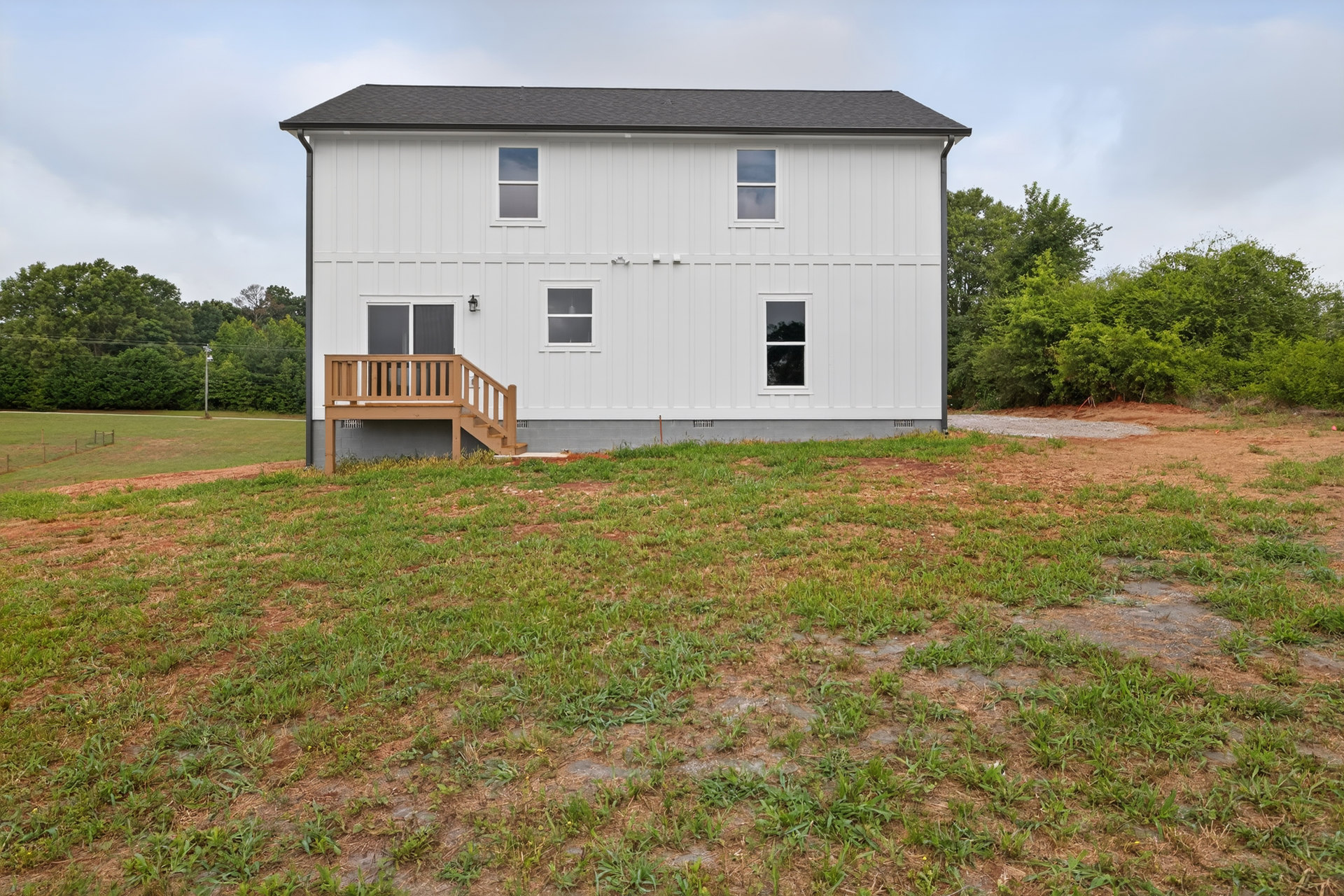 White farmhouse with wooden deck and staircase, surrounded by green grass lawn, white and grey exterior walls, large window with white frame, trees and cloudy sky in background