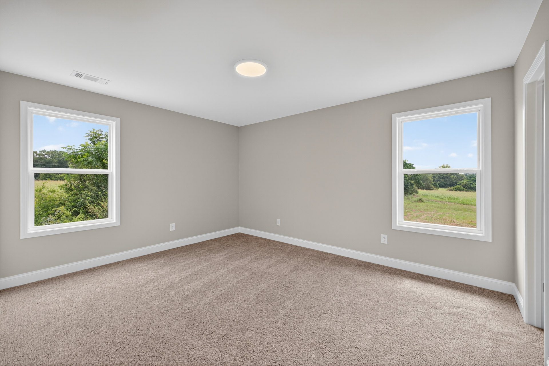 Carpeted room with two large windows, white walls, and views of trees and a grassy field outside