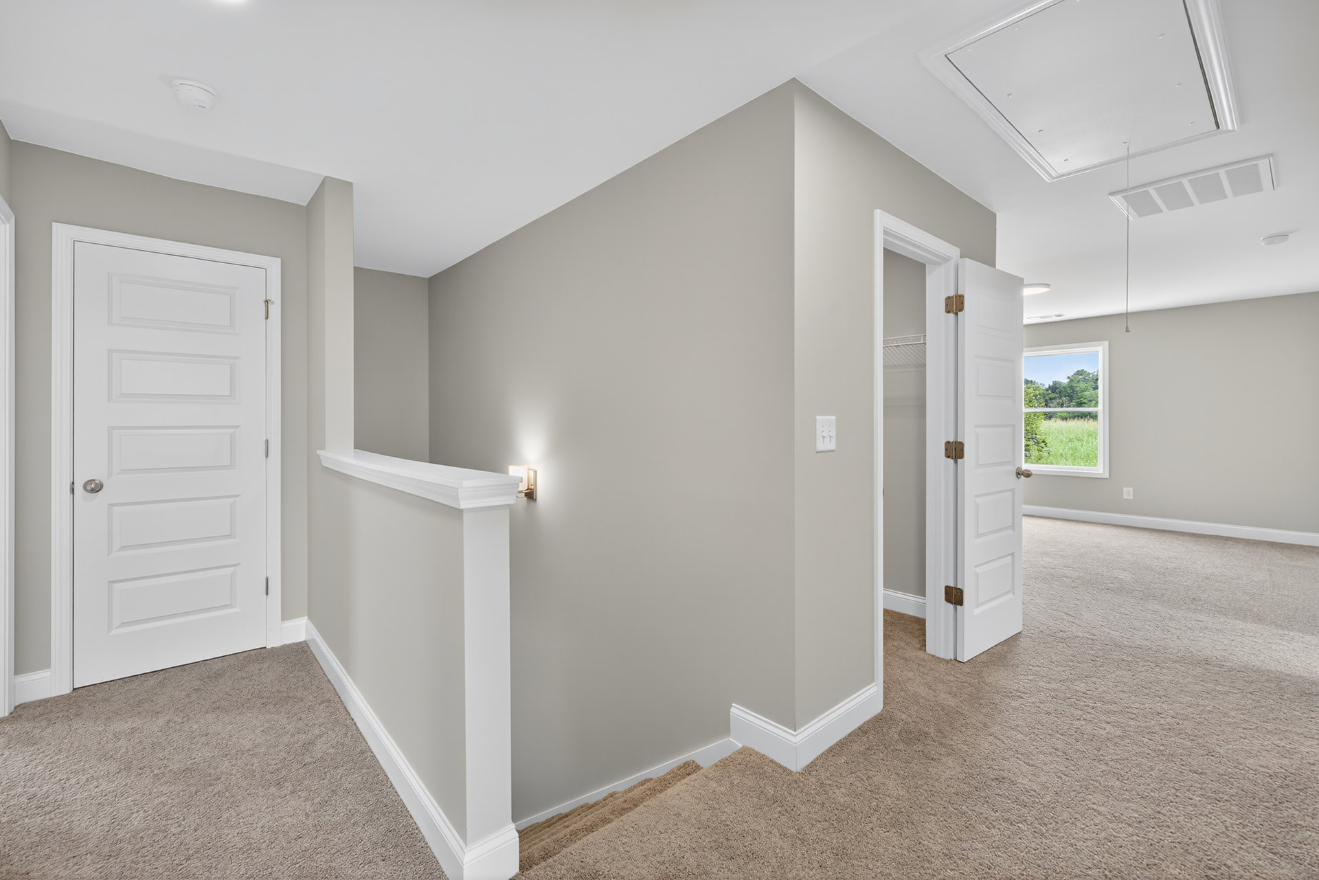 Hallway with white walls and carpeted floor, white door with silver handle, closet door, ceiling light fixture, window showing trees outside, ceiling with recessed holes