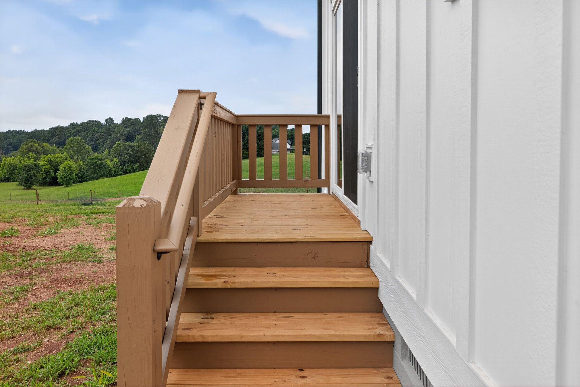 Wooden staircase leading up to a porch, surrounded by green grass and leafy trees, with a dirt patch and cloudy sky in the background