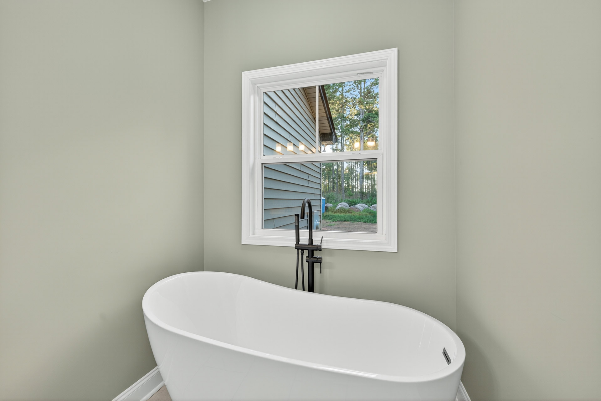 White freestanding bathtub beneath a window with outdoor views, black floor lamp and exposed black plumbing pipe, chrome faucet mounted on windowsill, light-colored walls
