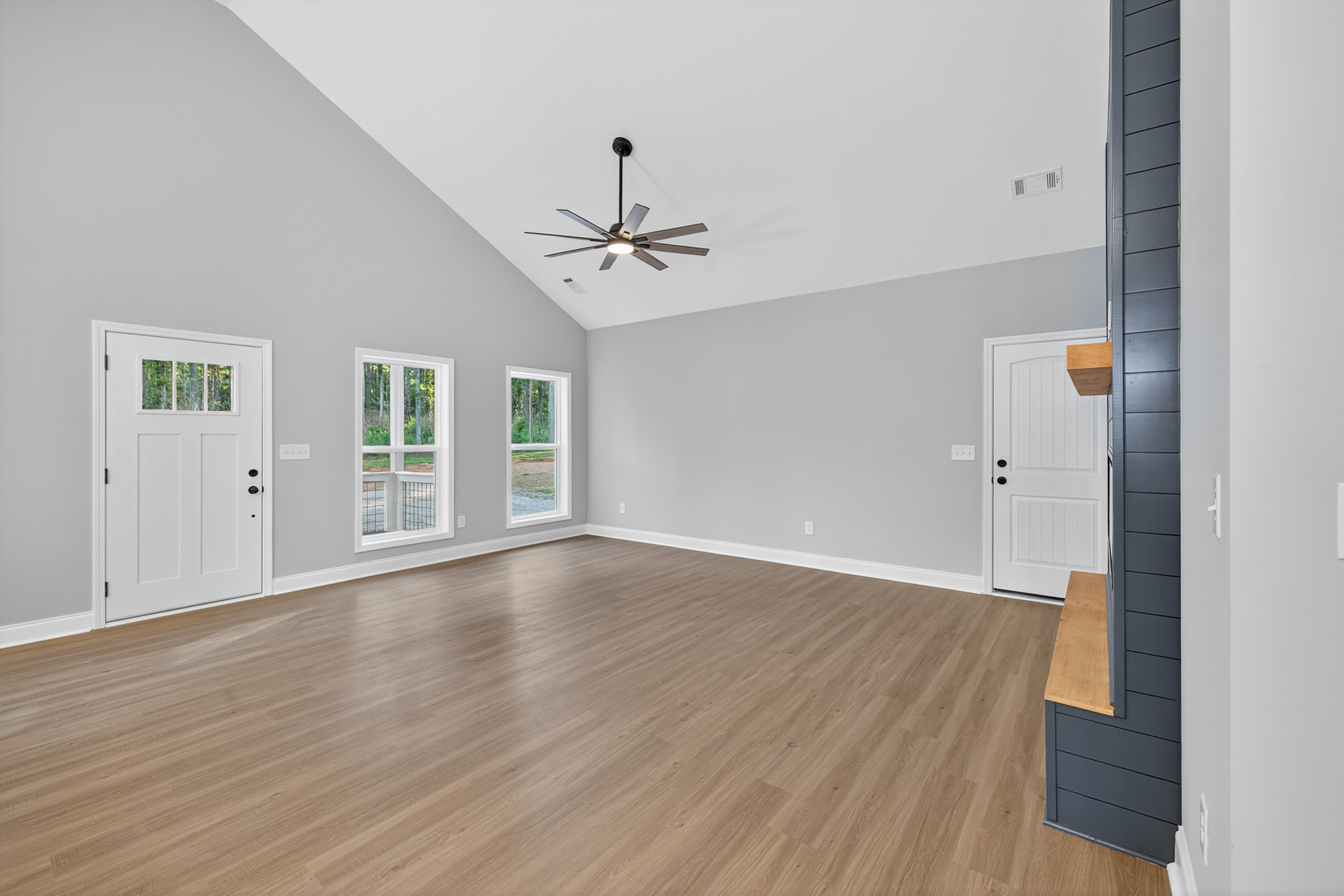 Ceiling fan with light fixture above wood flooring, white door with window and black knobs, large windows showing tree views, plaster walls