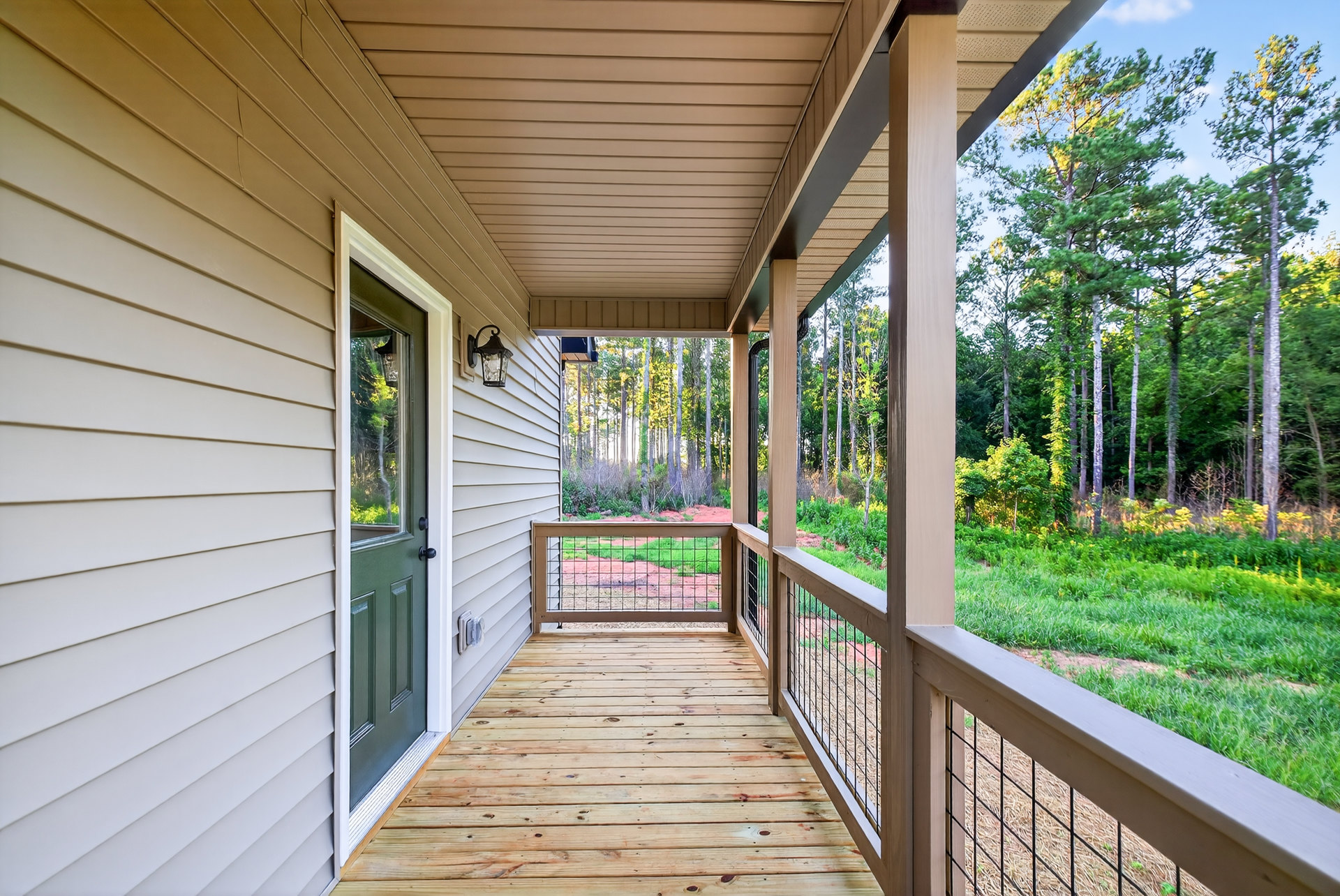 Green front door with window set in white siding, wooden porch deck with metal railing, fenced yard, and trees in the background.