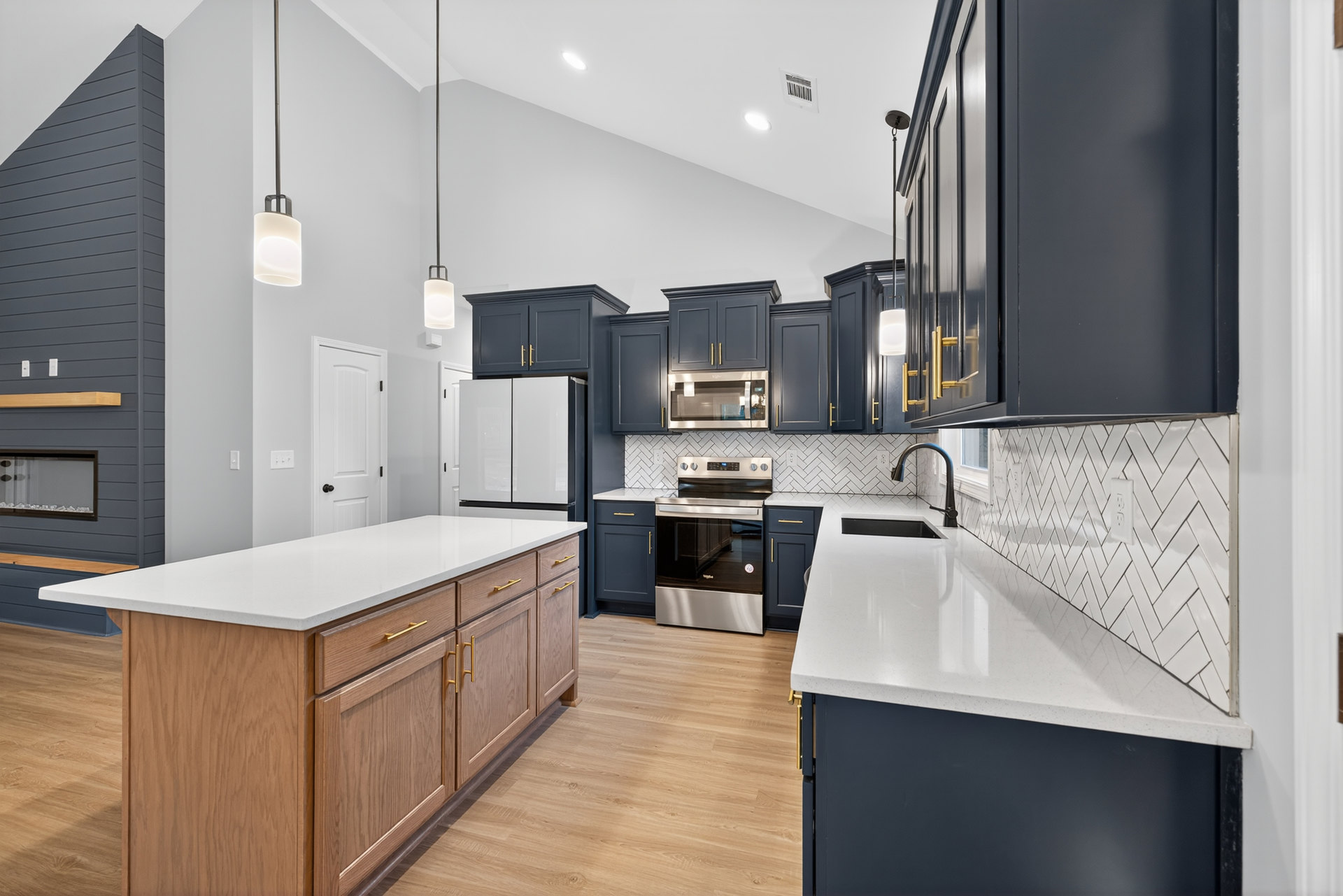 Blue kitchen cabinets with gold handles, white countertops, central island, stainless steel microwave and stove with glass door, tile backsplash, modern light fixture, undermount