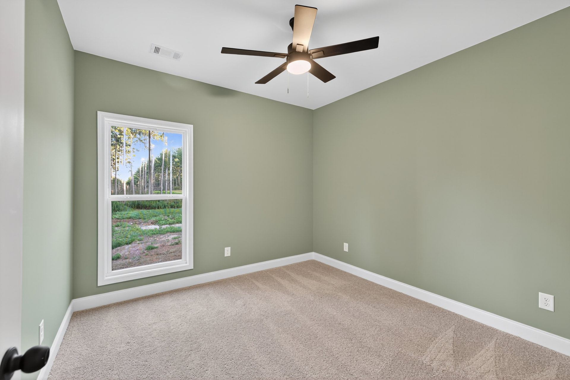 Bedroom with beige carpet, white walls, ceiling fan with light fixture, large window overlooking forest, white electrical outlet on wall