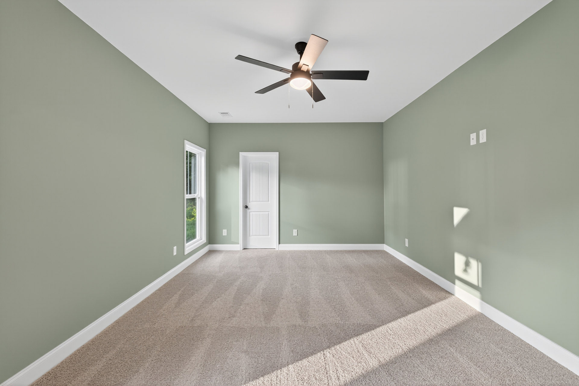 Carpeted bedroom with white walls, ceiling fan with light fixture, white door featuring black knob, and window with white trim.