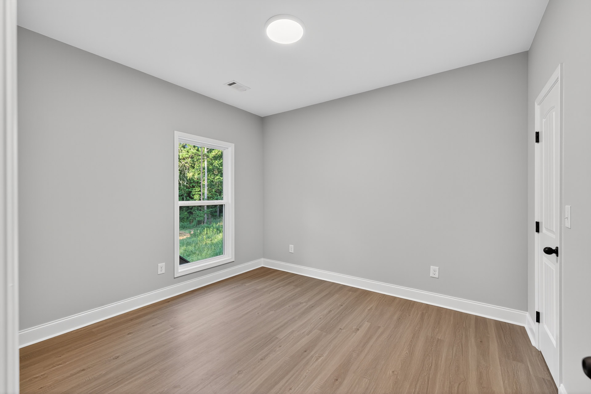 Sunlit room featuring wide-plank hardwood flooring, white plaster walls, large window overlooking forested landscape, and recessed ceiling light.
