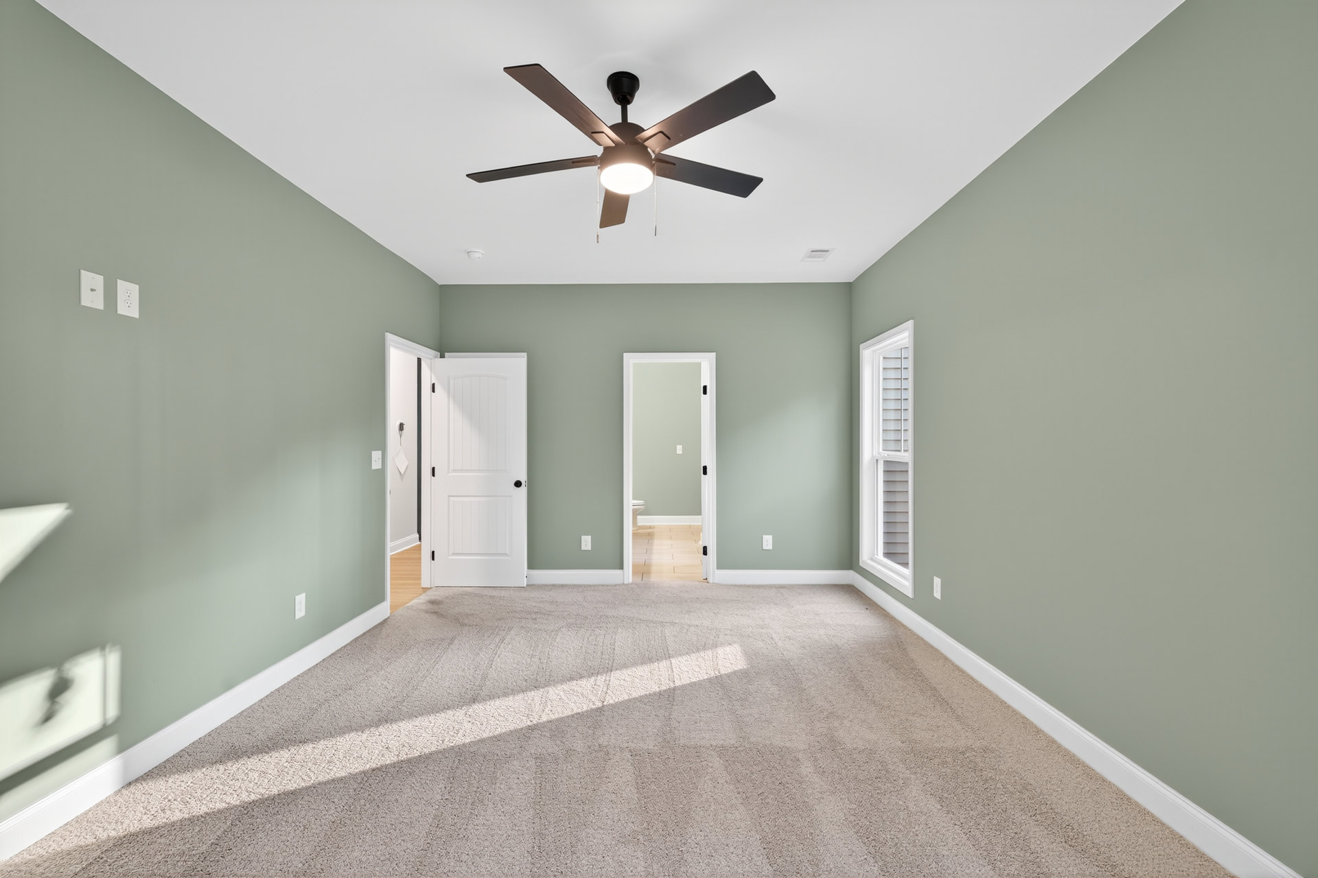 Bedroom with green accent wall, ceiling fan with light, white door featuring black hardware, window, and partial view of bathroom with toilet through open doorway