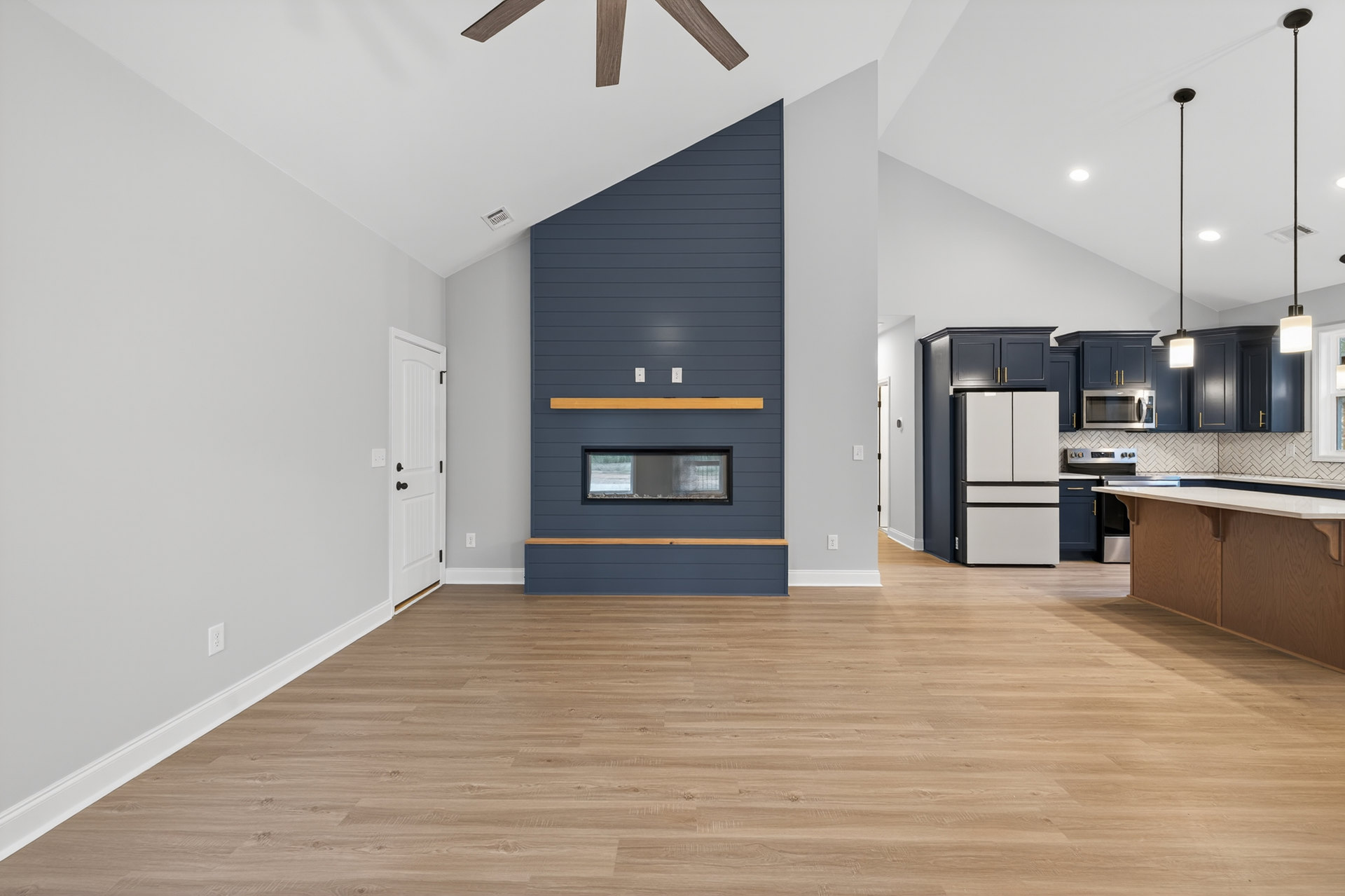Open-concept living area featuring a stone fireplace, adjacent kitchen with white cabinetry, black-trimmed refrigerator, glass window, light wood flooring, and blue accent wall