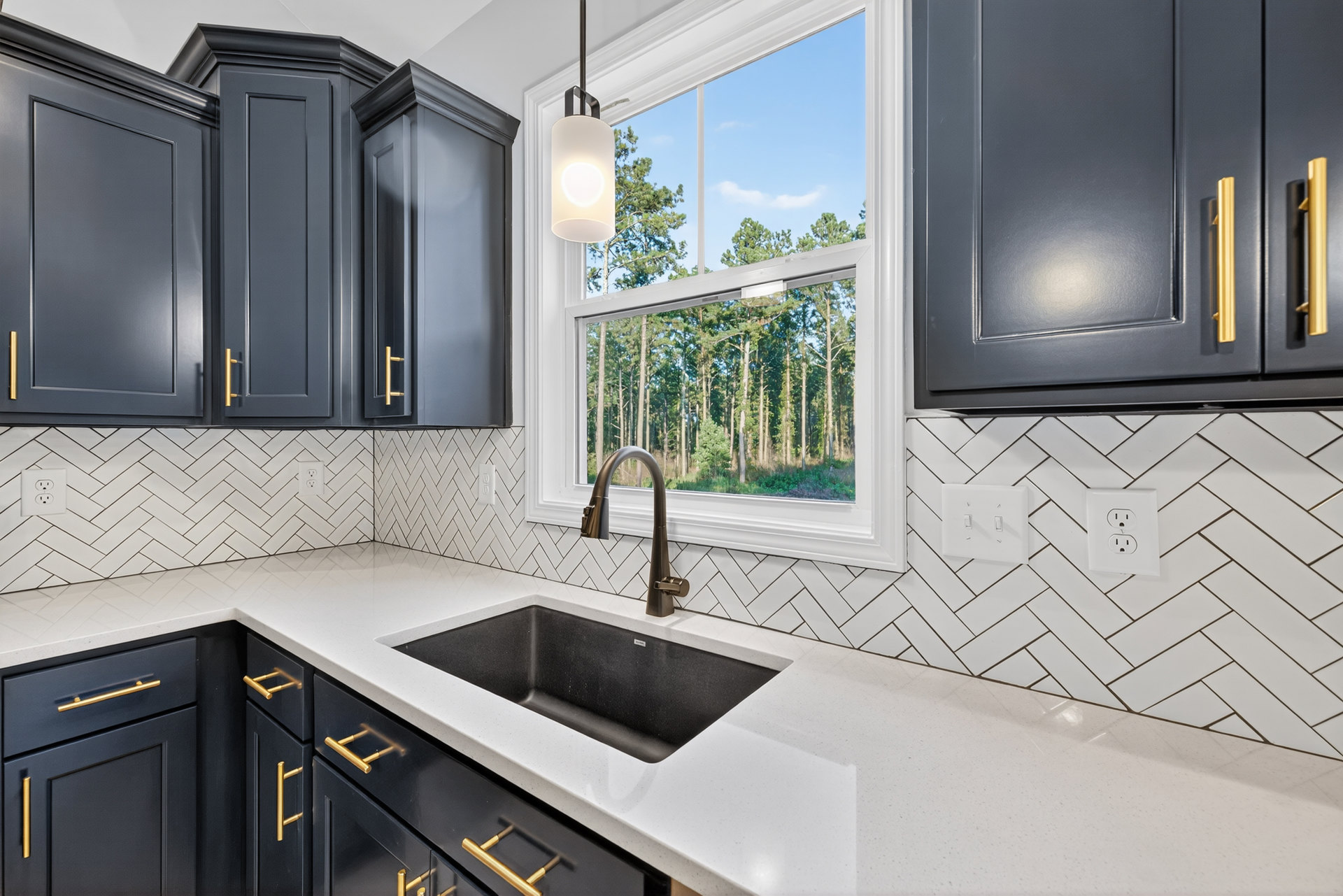 Modern kitchen featuring a stainless steel sink and faucet set against a white tile backsplash, light wood cabinets, white quartz countertop, window with green trees visible
