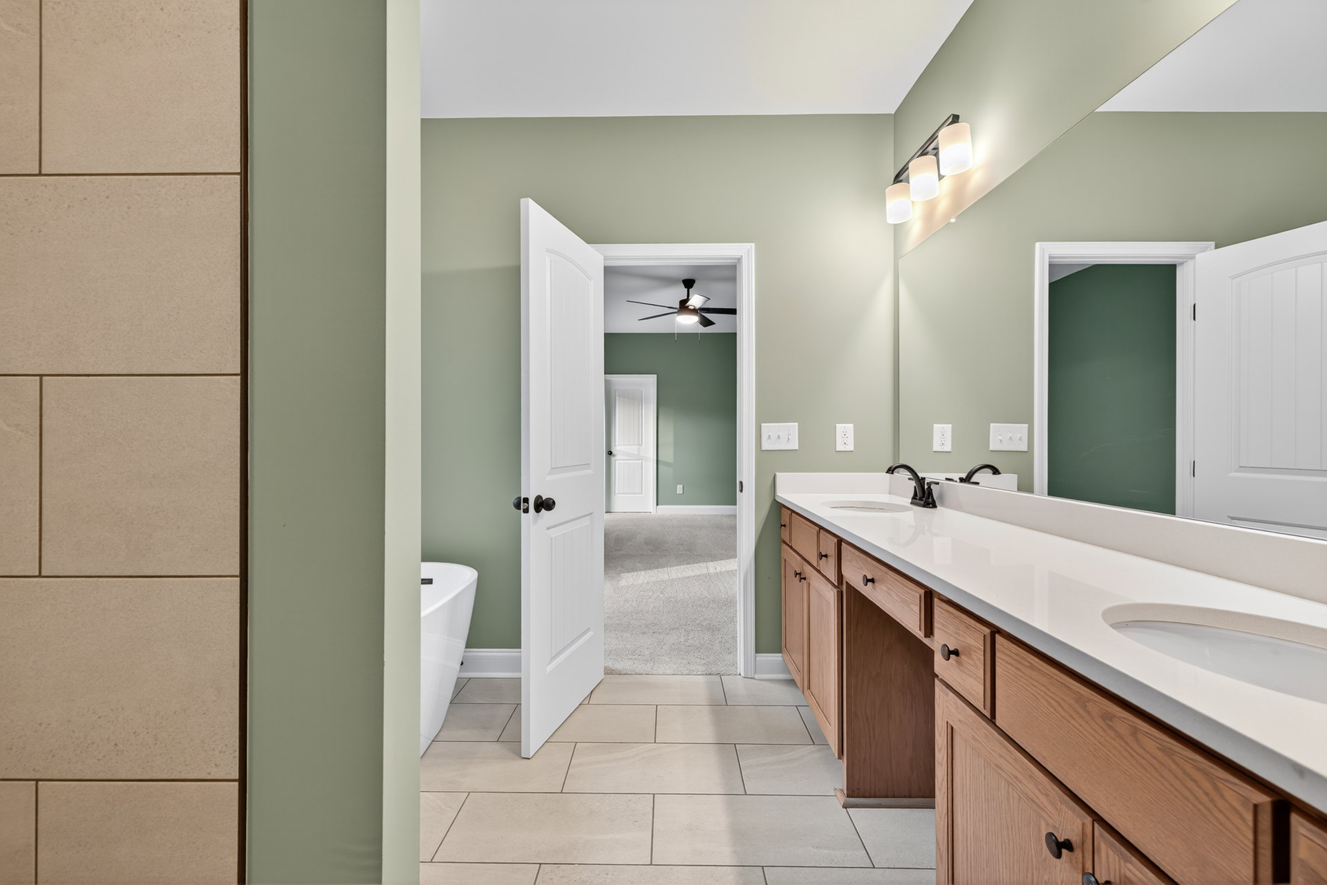 Bathroom with freestanding white bathtub, double sink vanity with stone countertop, white door featuring black handle, light tile flooring, and neutral wall finishes