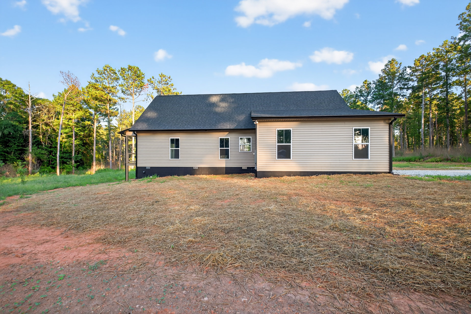 Single-story house with grey roof, white-framed windows, dirt yard, scattered trees, and blue sky with clouds
