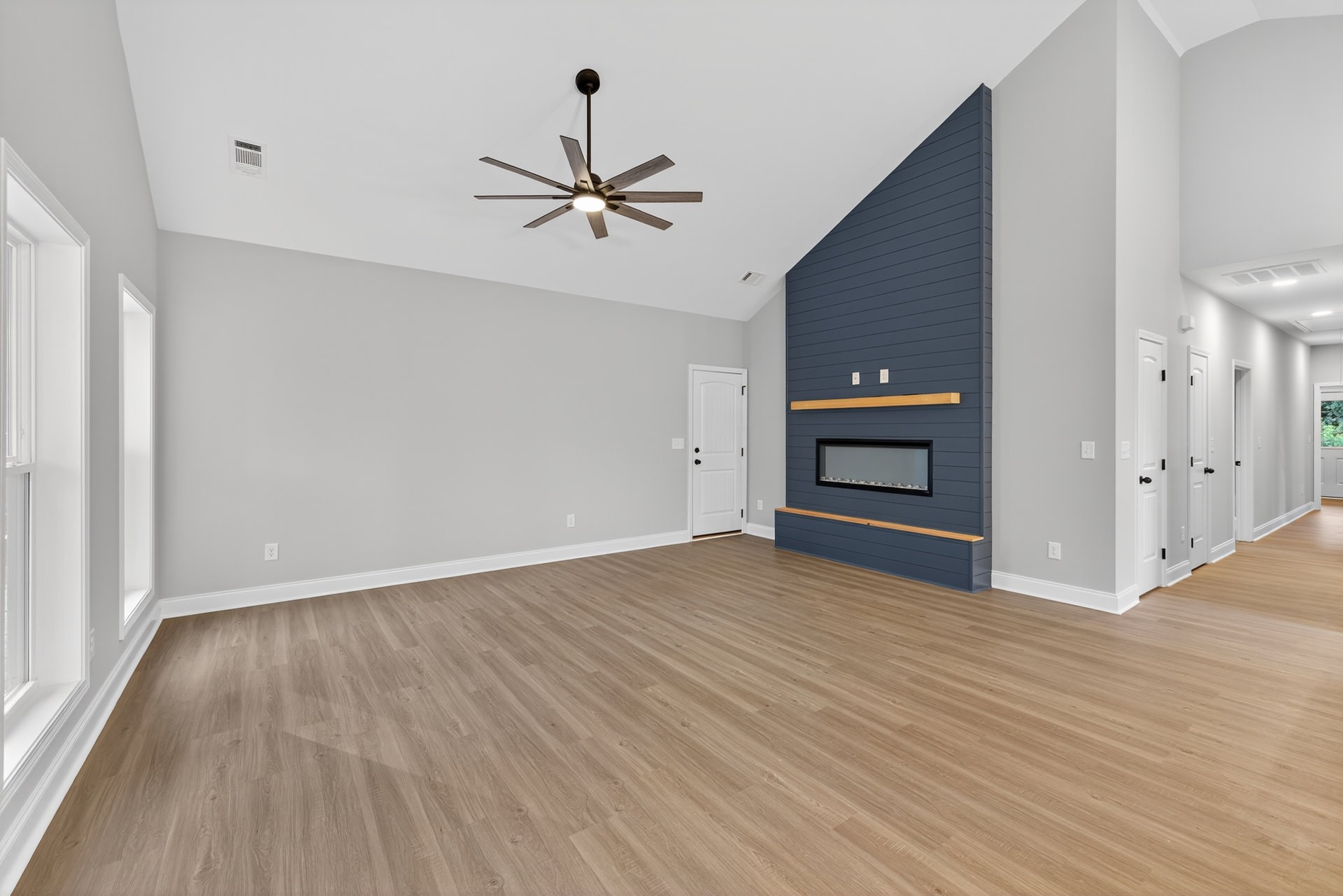 Living room with wood flooring, white walls, ceiling fan with light, fireplace with white surround, white door featuring black knobs, wall vent visible near baseboard