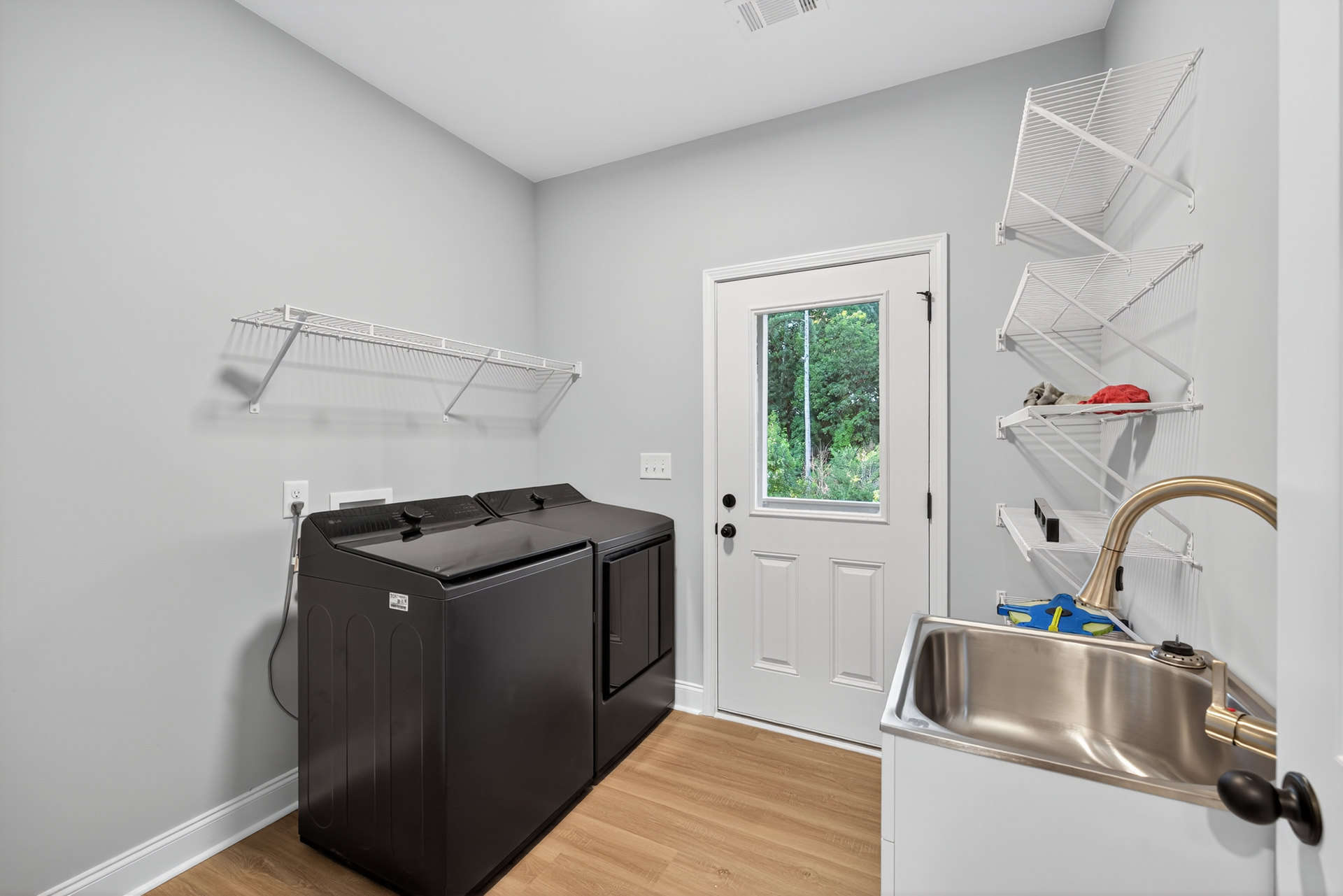 Laundry room with front-loading washer and dryer, white cabinetry, stainless steel sink with chrome faucet, white countertop, windowed door showing trees outside, metal shelving