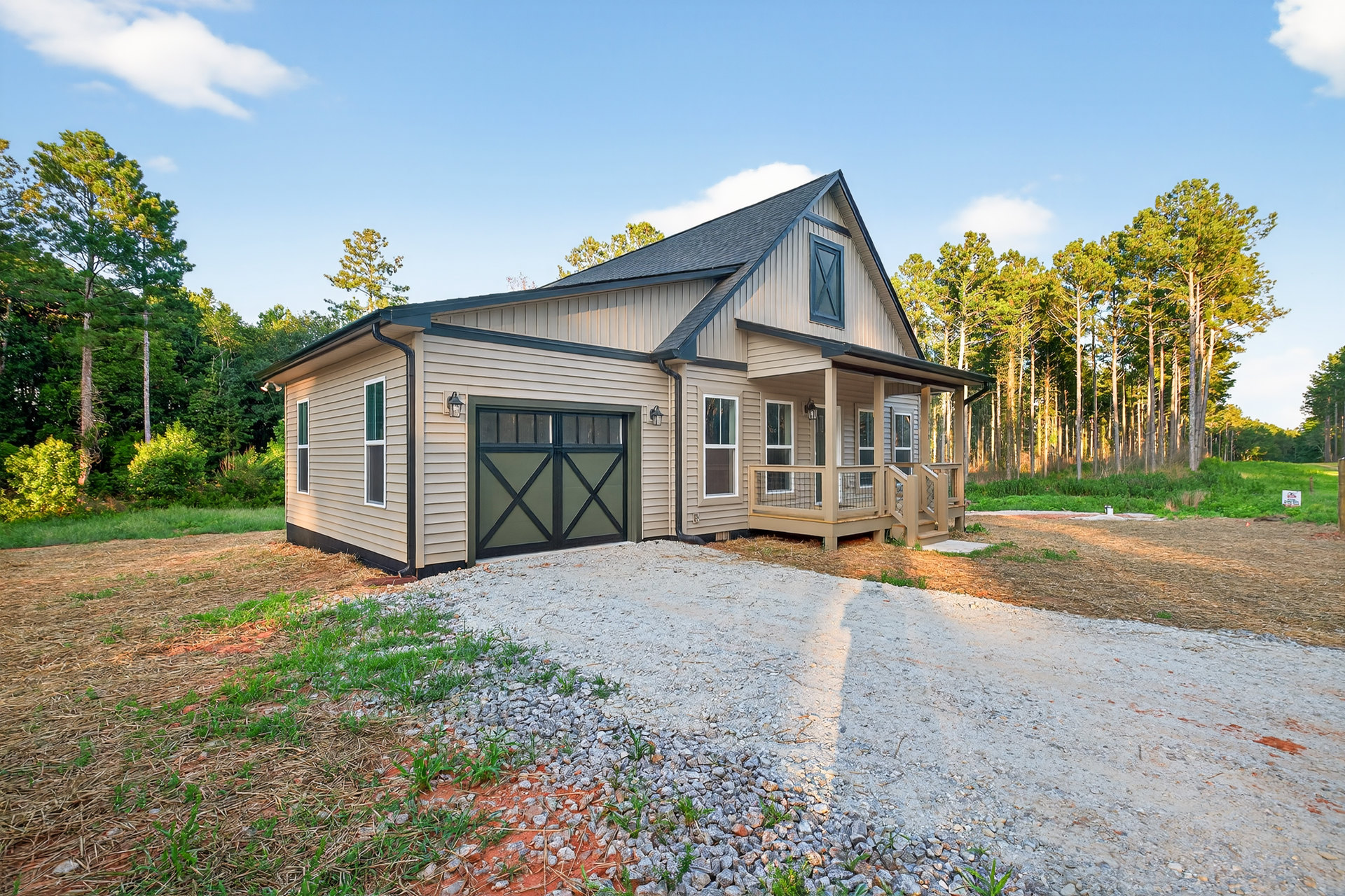 Two-story house with green garage door and black trim, gravel driveway, gray siding, covered porch, tall trees in background, manicured lawn