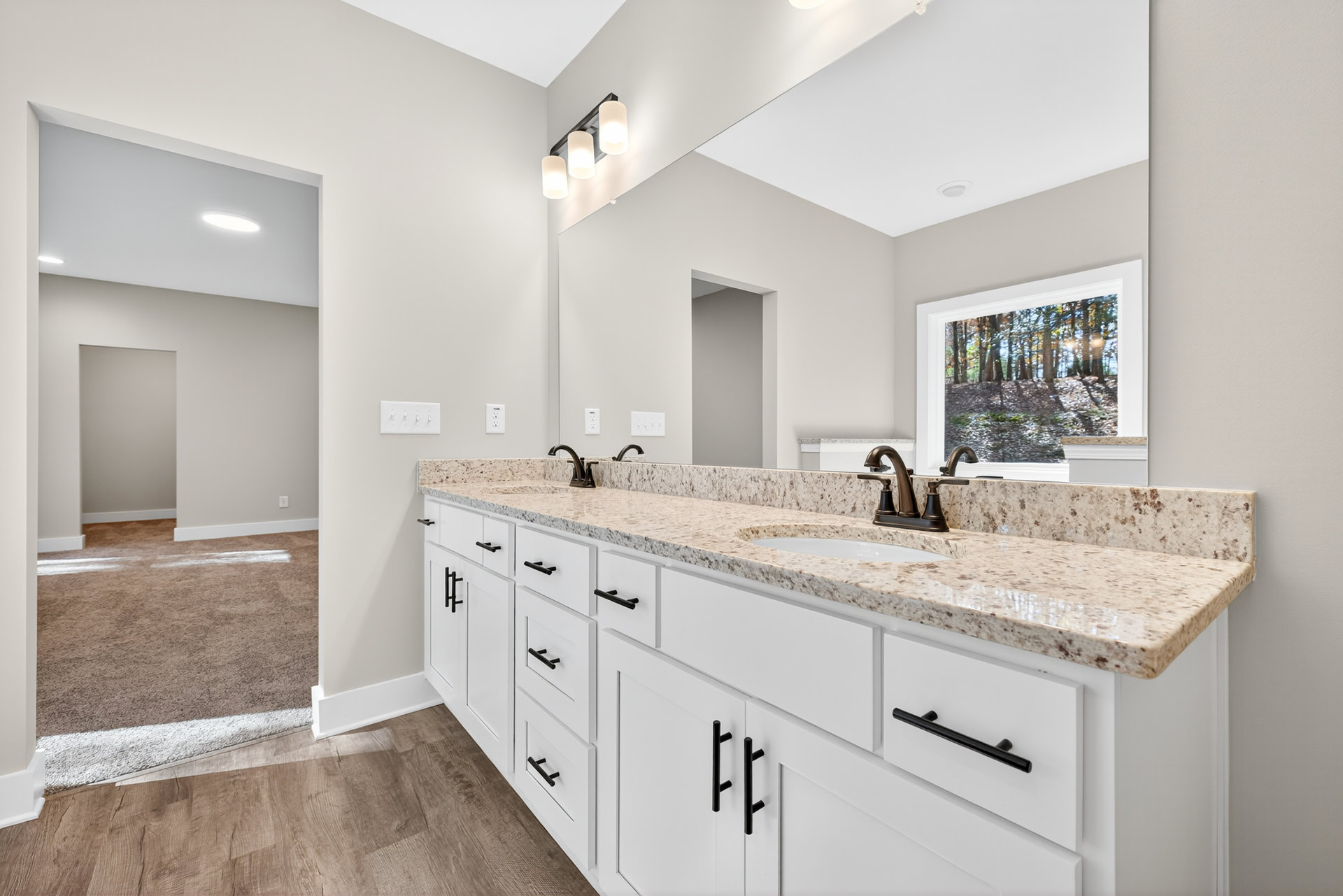 Bathroom with double sinks, marble countertop, white cabinets, large mirror, and window overlooking trees.