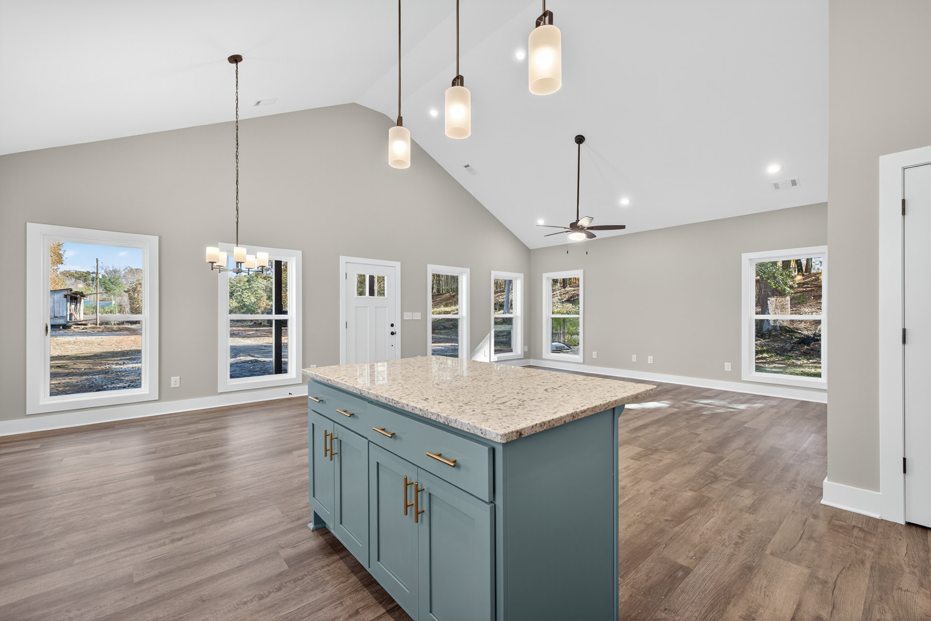 Open-concept kitchen featuring a marble-topped island, wood flooring, white cabinetry, pendant lighting, and large windows overlooking trees and a neighboring building.