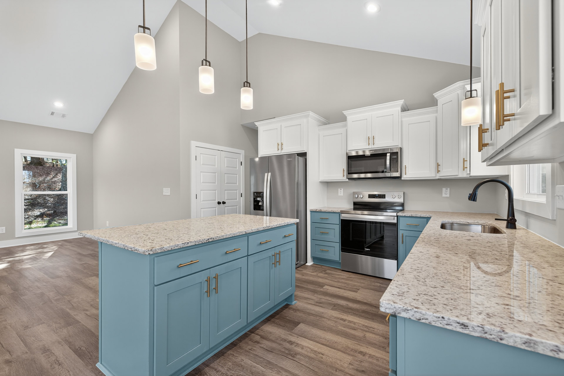 Kitchen featuring blue and white cabinets, marble-topped island, stainless steel stove and oven, built-in microwave, sink set in white countertop, window overlooking trees.