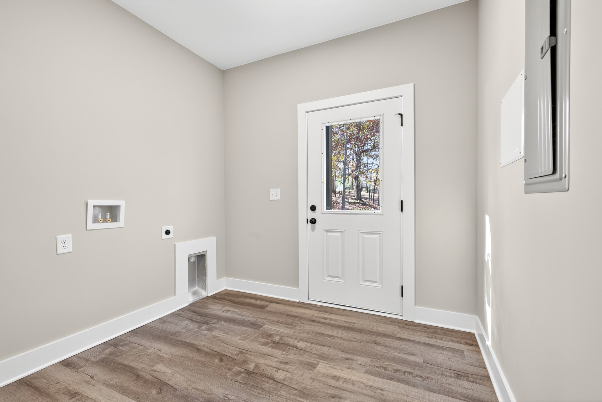 White door with glass window, wood laminate flooring, white baseboard trim, electrical outlet on plaster wall, window overlooking trees, small white storage box with objects