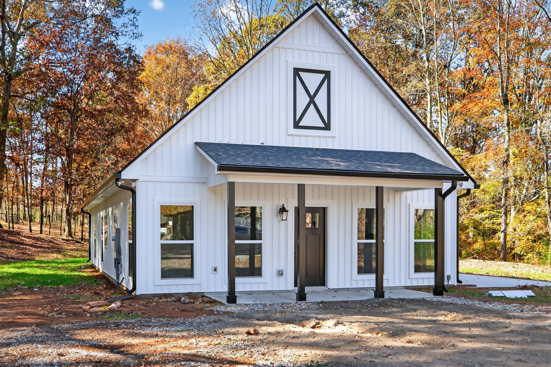White siding house with black shingle roof, brown front door, white-framed windows, dirt and rock landscaping, autumn trees in background